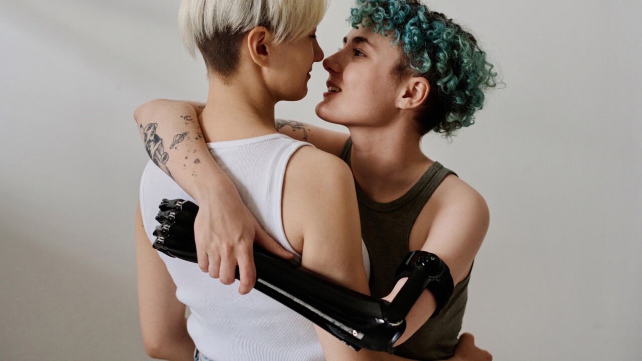 Auf dem Bild sind zwei Frauen zu sehen, die sich umarmen und sich gegenseitig tief in die Augen schauen. Die linke Frau hat blonde Haare, die rechte Frau hat gründe Haare. Die rechte Frau trägt eine Handprothese am linken Arm.