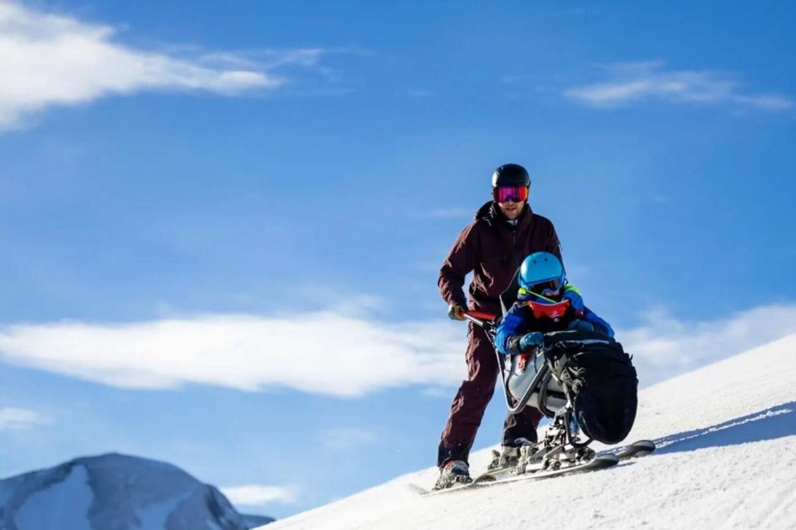 Eine Person hilft einem Kind in einer adaptiven Skiausrüstung einen verschneiten Abhang hinunter, beide tragen Helme und Schutzbrillen, im Hintergrund sind ein blauer Himmel und Berge zu sehen.