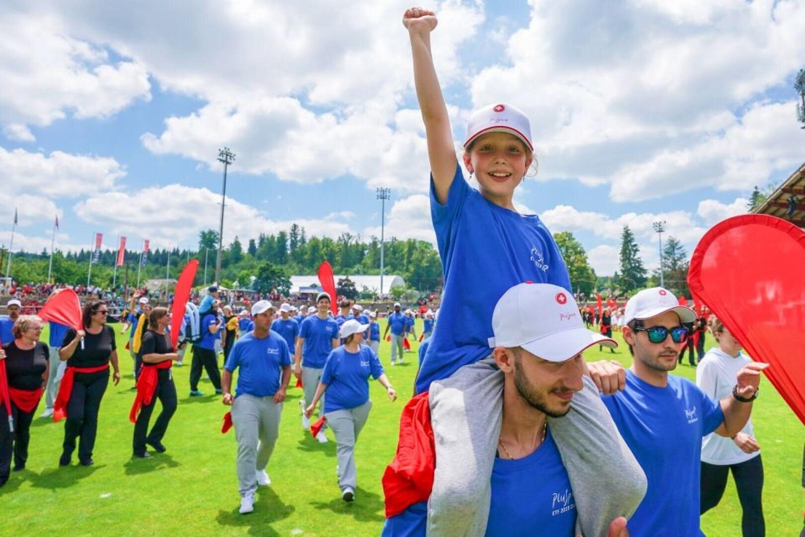 Ein lächelndes Kind in einem blauen Hemd und einer weißen Mütze hebt eine Faust, während es auf den Schultern eines Mannes in einer Menschenmenge bei einer Veranstaltung im Freien sitzt. Viele Menschen in passenden blauen Hemden und roten Requisiten umgeben sie an einem sonnigen Tag.