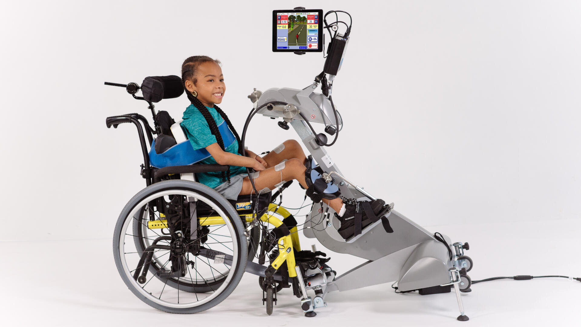 Ein lächelndes Kind im Rollstuhl benutzt ein robotisches Rehabilitationsgerät für Beinübungen, auf dessen Bildschirm ein Spiel zur interaktiven Therapie angezeigt wird. Das Kind hat Elektroden an den Beinen und ist mit der Aktivität beschäftigt.