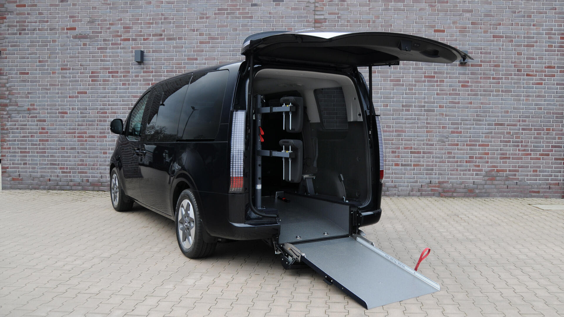 Ein schwarzer Lieferwagen mit geöffneter Heckklappe und einer Rollstuhlrampe, die auf eine gepflasterte Fläche ausgefahren ist, parkt vor einer Backsteinmauer. Das Innere des Lieferwagens ist durch die offene Heckklappe sichtbar.