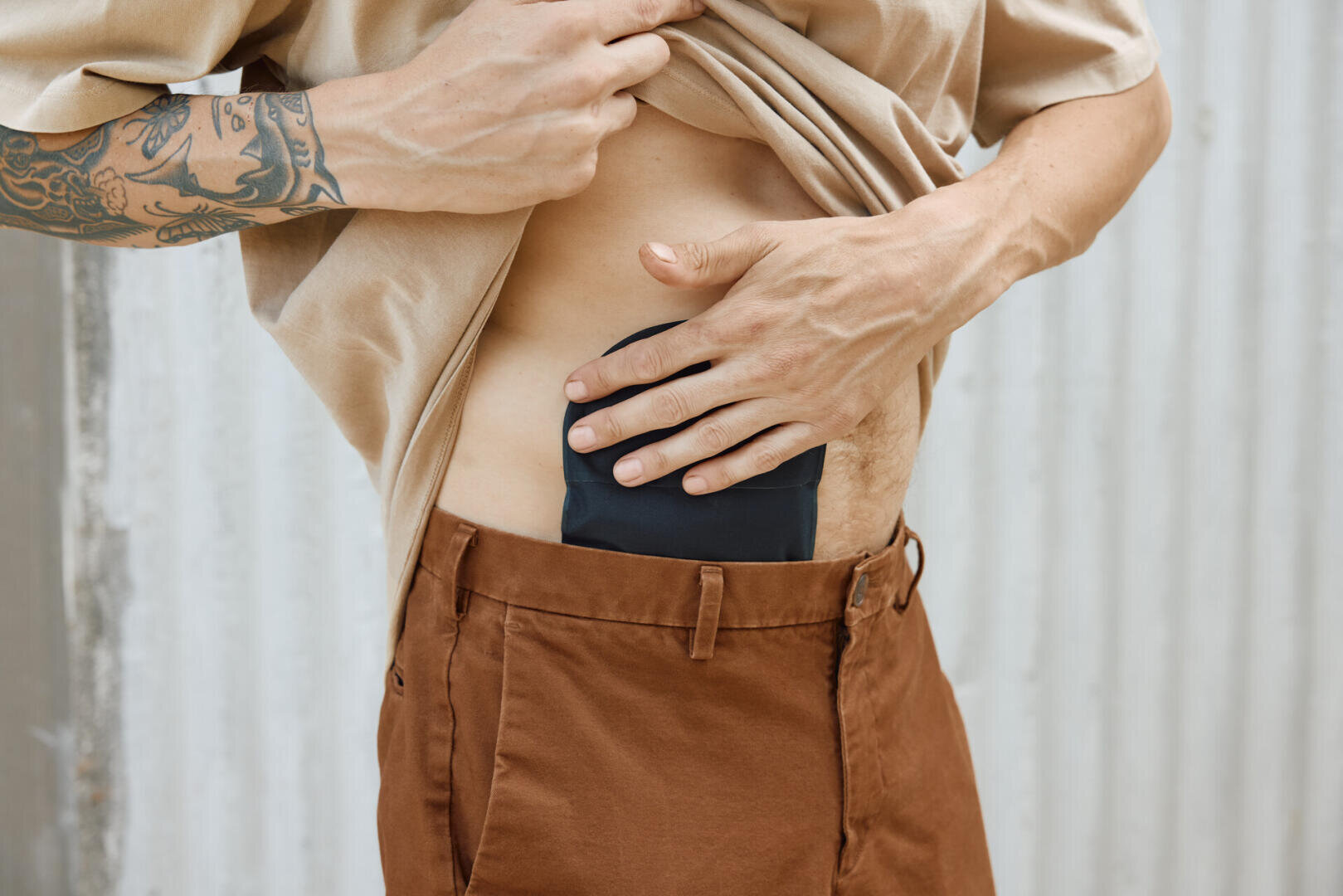 Eine Person mit einem tätowierten Arm hebt ihr Hemd und zeigt einen am Bauch befestigten Stomabeutel über einer braunen Hose, die vor einem hellen, gewellten Hintergrund steht.