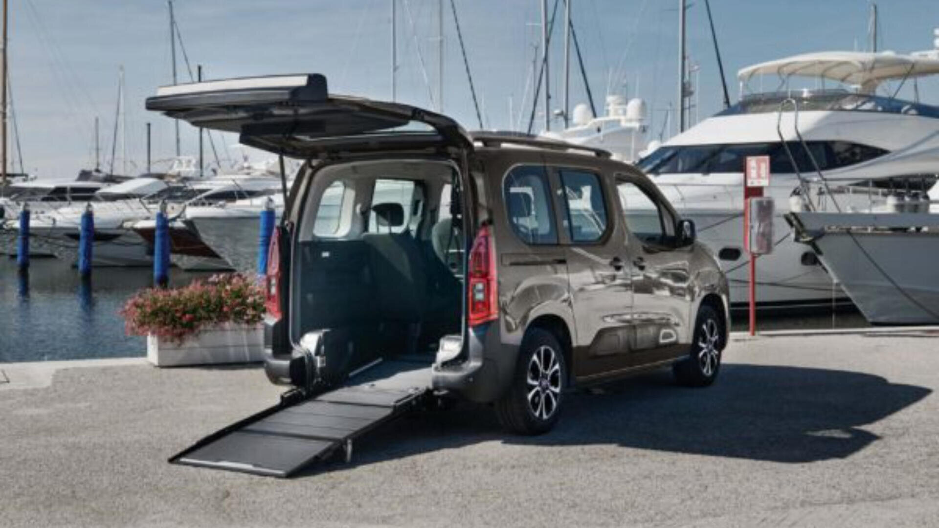 Ein silberner Lieferwagen mit geöffneter Heckklappe und ausgefahrener Rollstuhlrampe parkt an einem Yachthafen, im Hintergrund sind mehrere Yachten und Boote zu sehen.