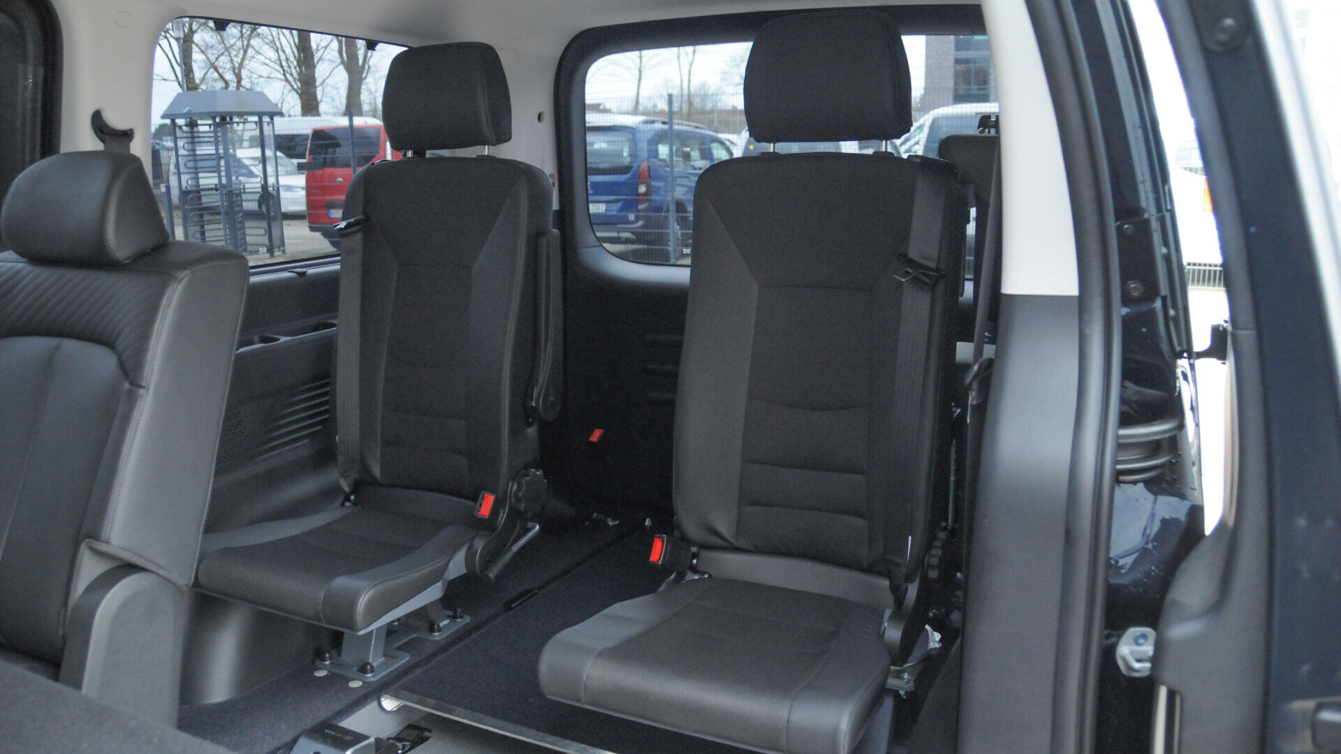 Zwei schwarze Autositze befinden sich im hinteren Teil eines Lieferwagens, wobei eine große Seitentür geöffnet ist. Die Sitze sind getrennt und aufrecht, und zwischen ihnen und den Vordersitzen ist reichlich Platz. Draußen sind ein Fenster und geparkte Autos zu sehen.