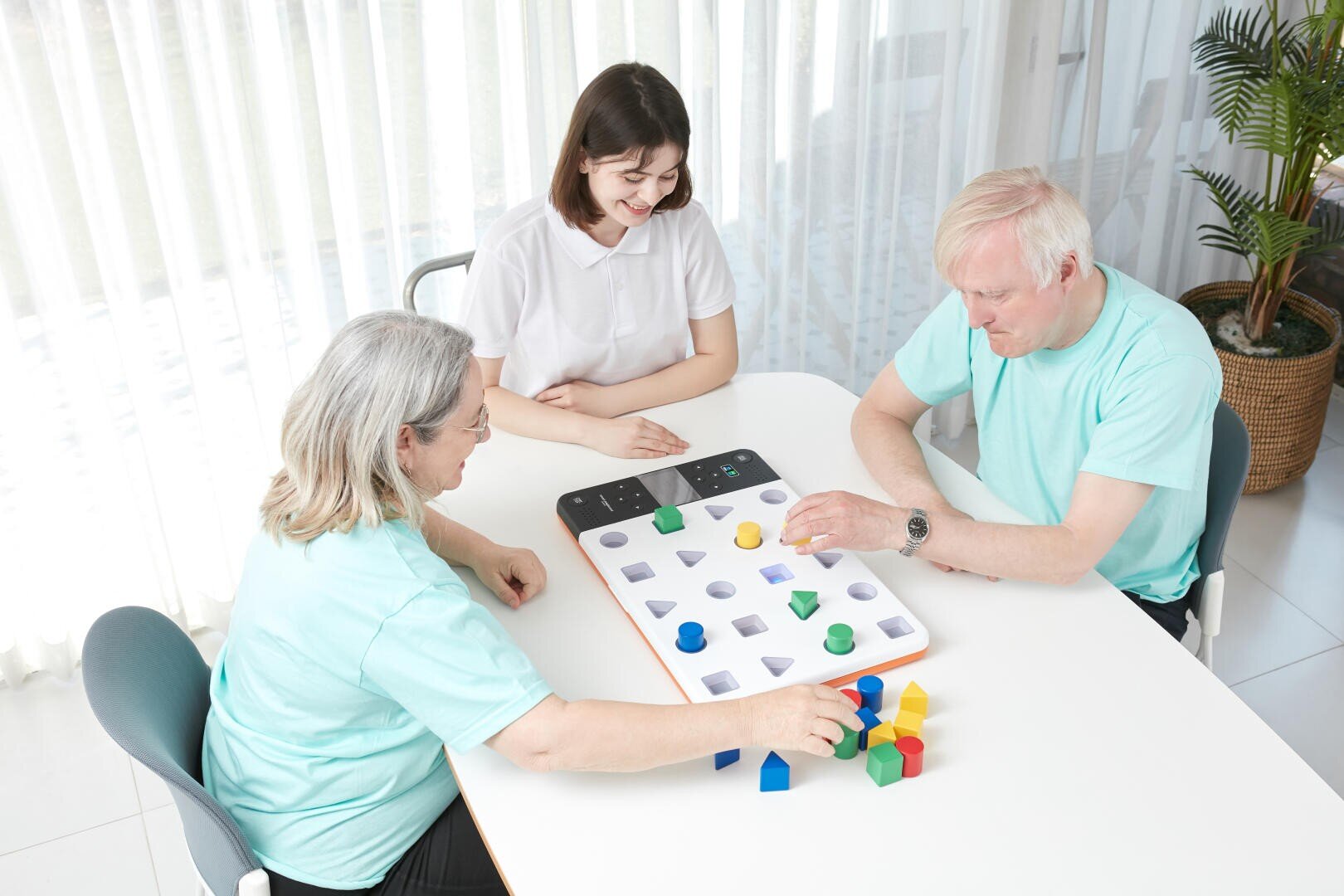 Zwei ältere Erwachsene und eine jüngere Frau sitzen an einem Tisch und spielen ein Spiel mit bunten geometrischen Formen, die sie den entsprechenden Feldern auf einem Brett zuordnen. Die Gruppe scheint konzentriert und engagiert bei der Sache zu sein.