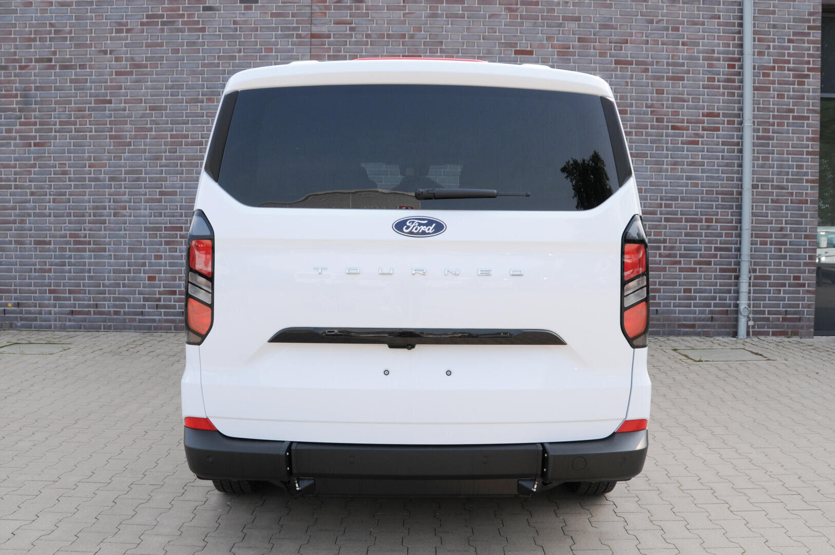 Ein weißer Ford Tourneo Transporter ist auf einer gepflasterten Fläche vor einer Backsteinmauer geparkt, direkt von hinten gesehen.