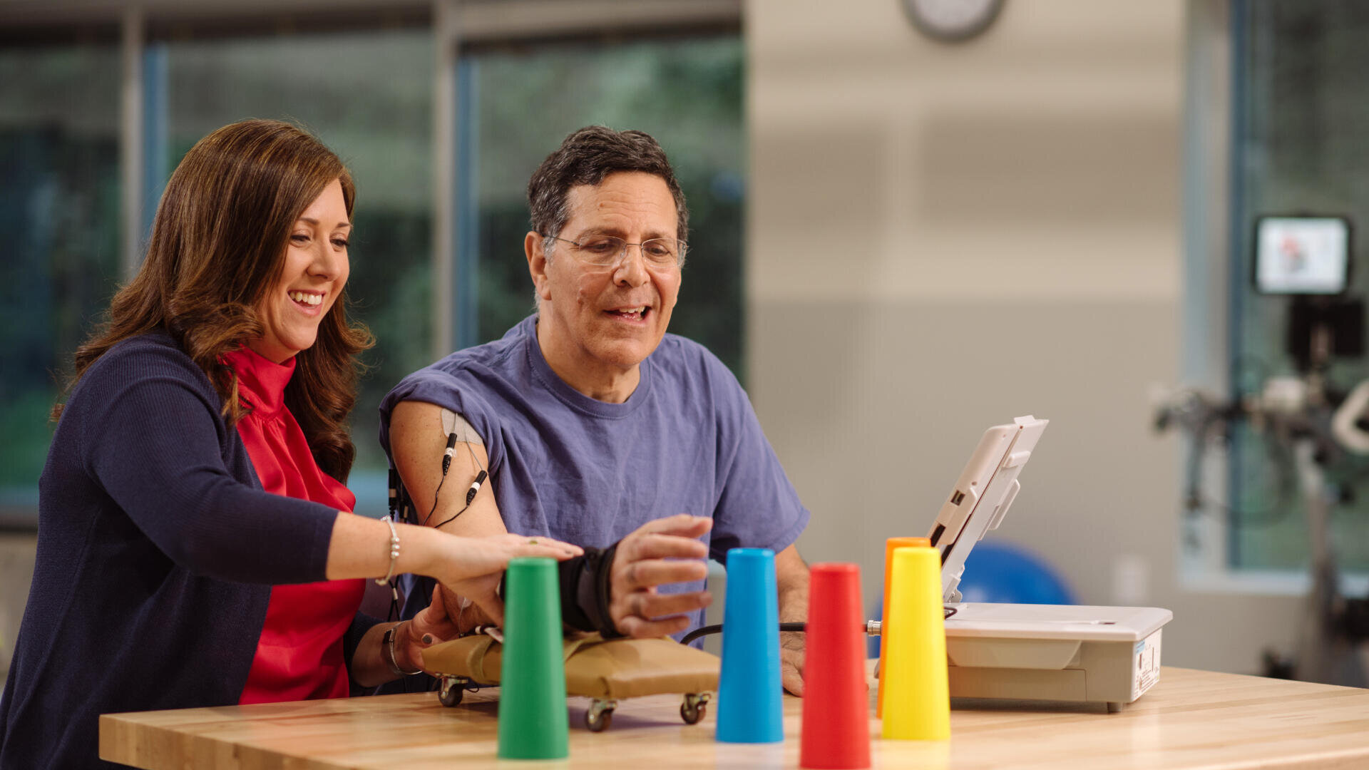 Eine Frau hilft einem Mann mit einer Armprothese, während er bunte Plastikbecher auf einem Tisch in einem hellen Raum stapelt, was auf eine Rehabilitations- oder Therapiesitzung hindeutet.