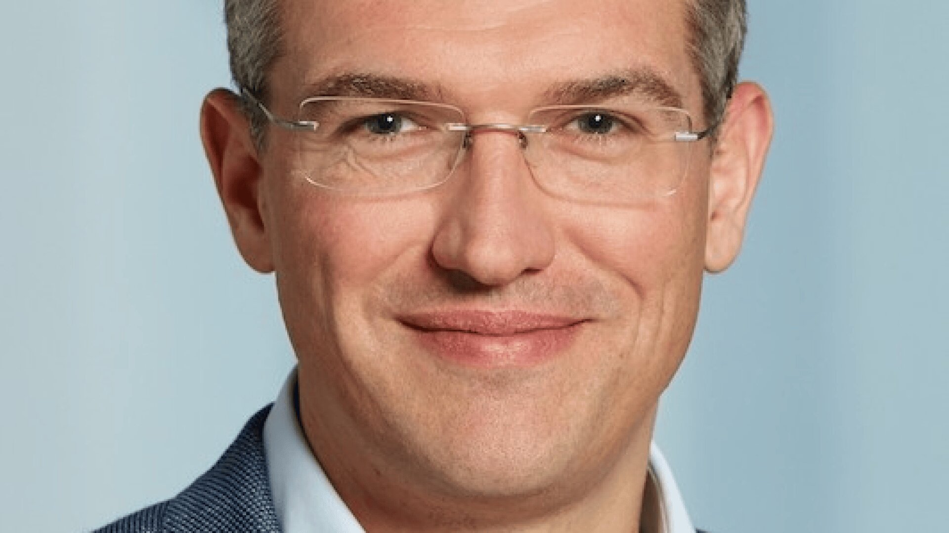 Ein Mann mit kurzen hellbraunen Haaren, der eine Brille und einen blauen Blazer trägt, lächelt vor einem hellblauen Hintergrund in die Kamera.
