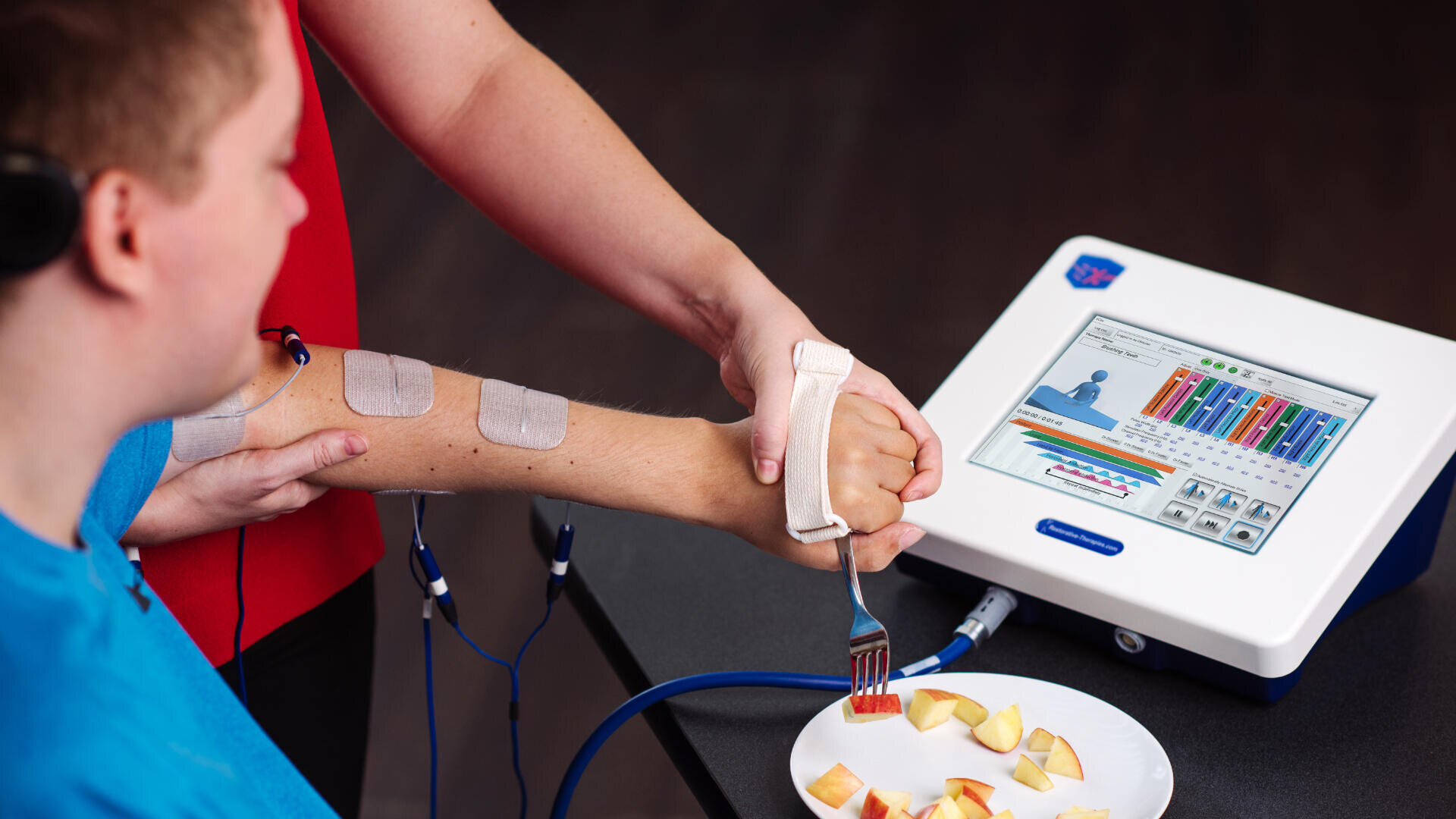 Eine Person hilft einer anderen Person mit Elektroden am Arm, mit einer Gabel Apfelscheiben von einem Teller zu nehmen, während auf einem Bildschirm in der Nähe ein medizinisches Gerät mit Tabellen und Diagrammen angezeigt wird.