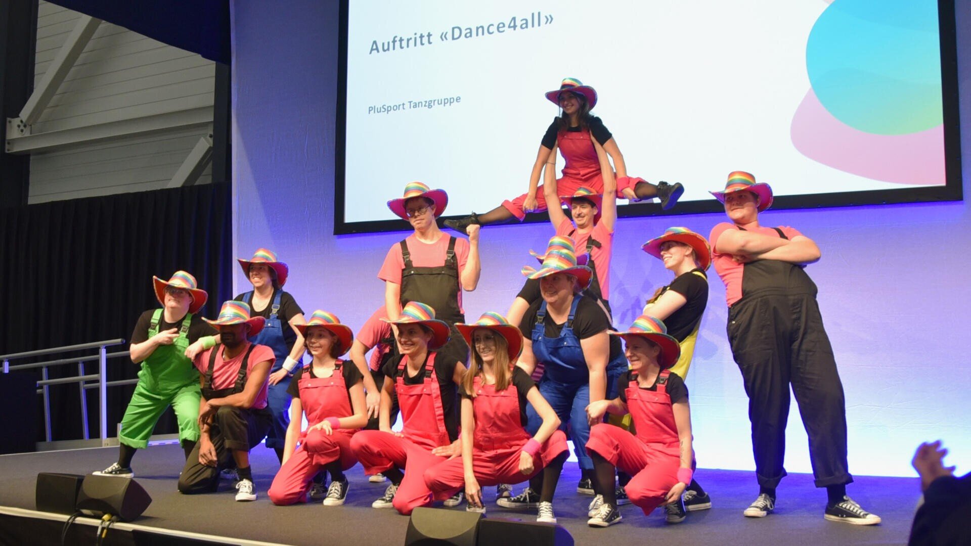 Eine Gruppe von Darstellern mit bunten Hüten und Overalls posiert auf der Bühne, einige stehen, einige knien, und eine Person balanciert auf den anderen. Auf einer Leinwand hinter ihnen ist der Text zu lesen: Auftritt "Dance4all" und PluSport Tanzgruppe.