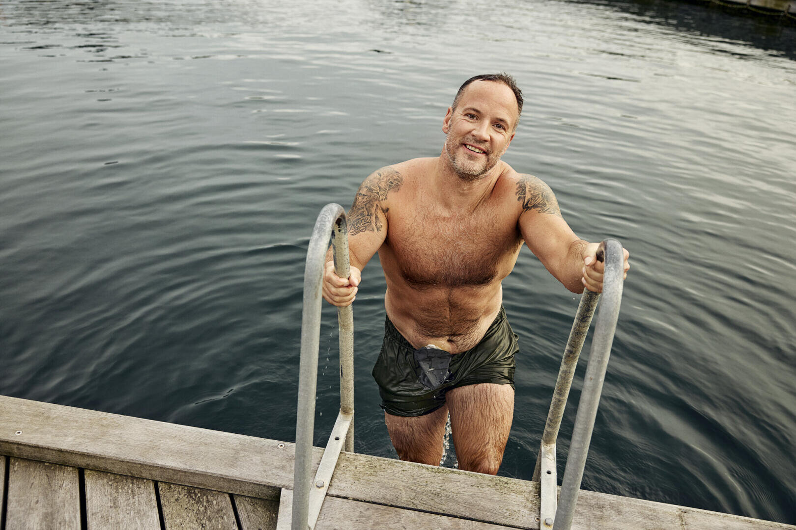 Ein lächelnder Mann mit Tattoos klettert in grünen Badehosen eine Metallleiter aus dem Wasser hinauf auf einen Holzsteg.
