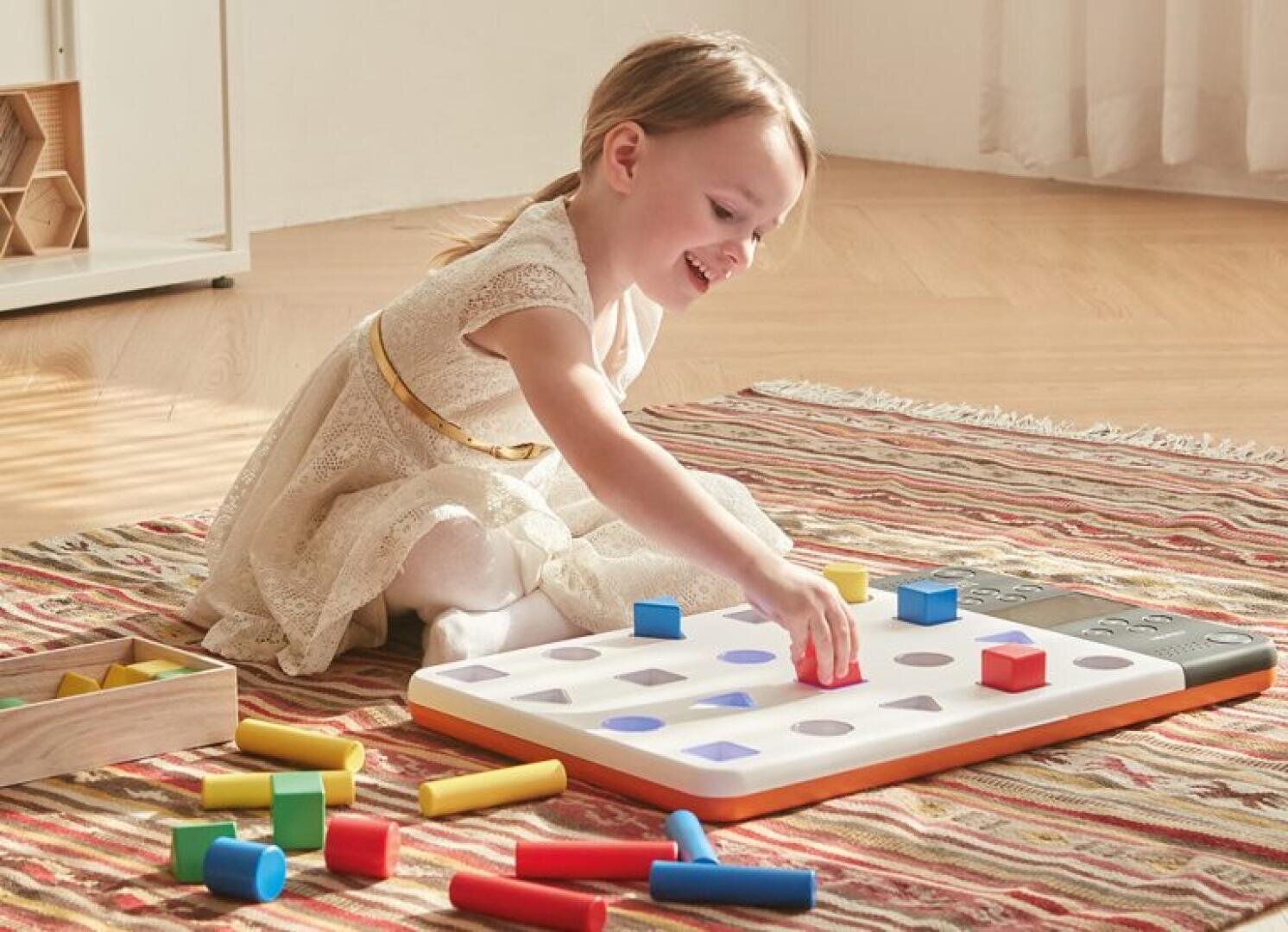 Ein kleines Kind in einem weißen Kleid sitzt auf einem gemusterten Teppich und lächelt, während es mit einem Spielzeug zum Sortieren von Formen spielt und bunte Blöcke in passende Löcher auf einer weißen Tafel steckt.