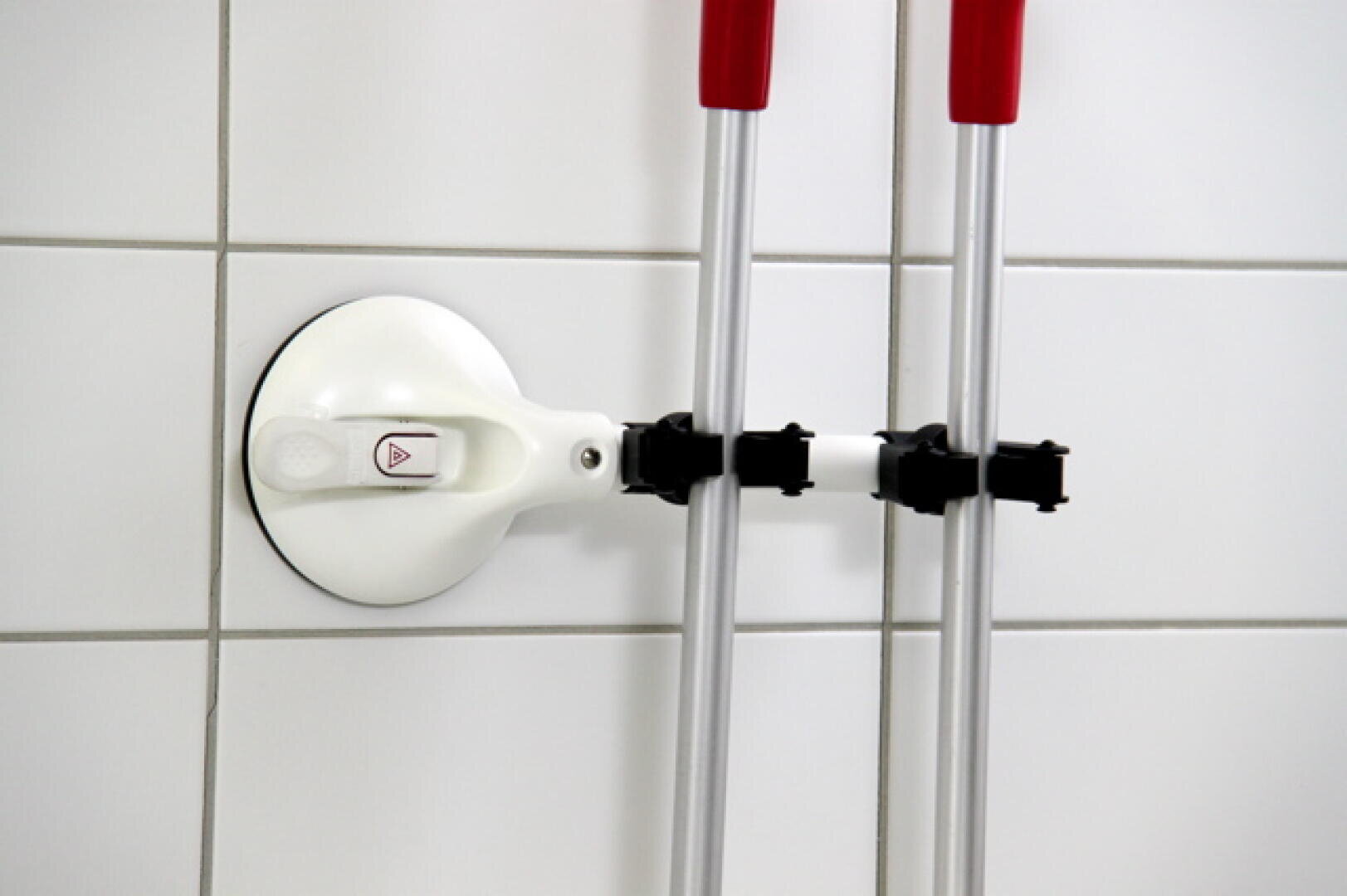 Ein weißer Wandhalter mit schwarzen Clips hält zwei Reinigungswerkzeugstiele mit roter Spitze an einer gefliesten Wand fest.
