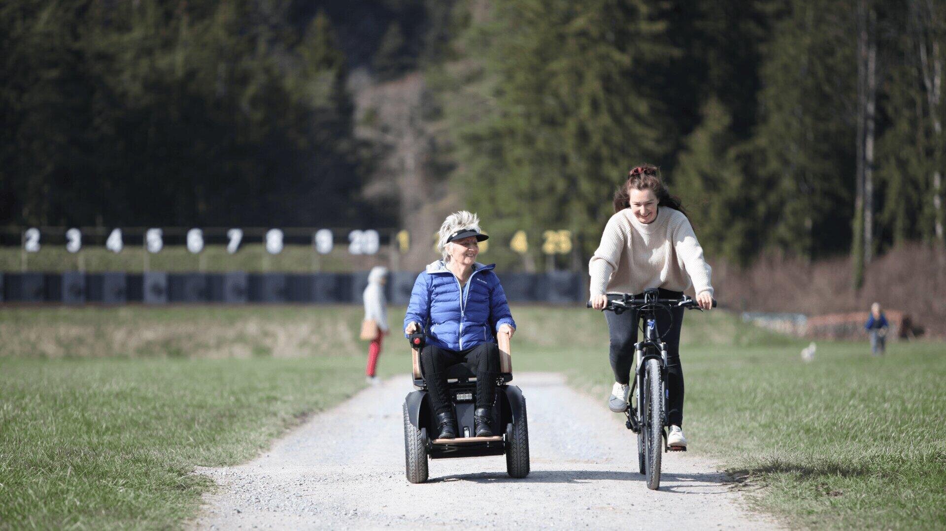 Eine ältere Frau in einer blauen Jacke fährt mit einem Elektroroller auf einem Weg neben einer jungen Frau auf einem Fahrrad. Sie befinden sich im Freien auf einer Wiese mit Bäumen, lächeln und genießen den Tag zusammen.