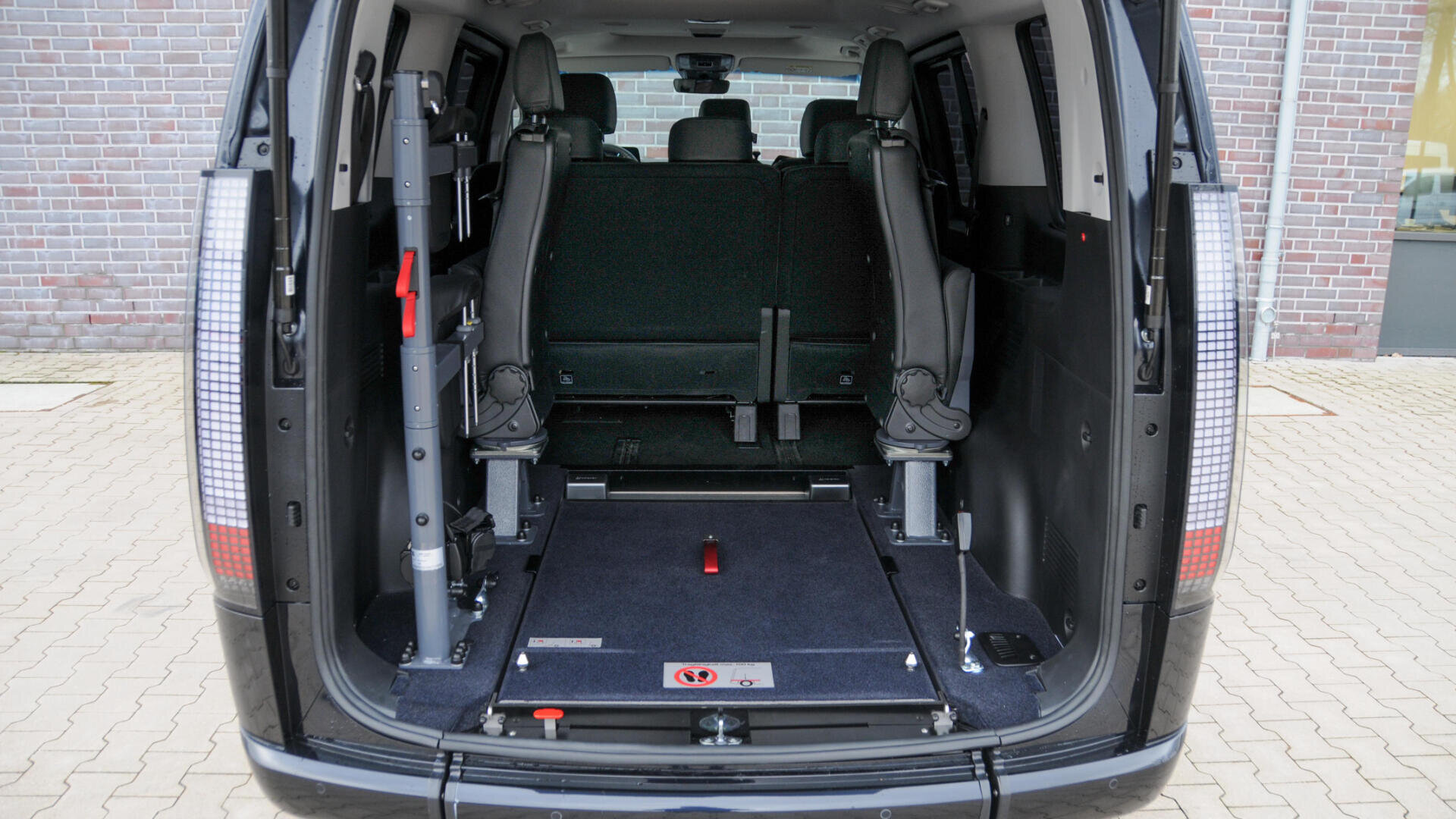 Innenansicht eines rollstuhlgerechten Lieferwagens mit umgeklappten Sitzen an den Seiten und einem leeren Raum mit abgesenktem Boden für den Rollstuhl. Der Lieferwagen ist auf einer gepflasterten Fläche in der Nähe einer Backsteinmauer geparkt.