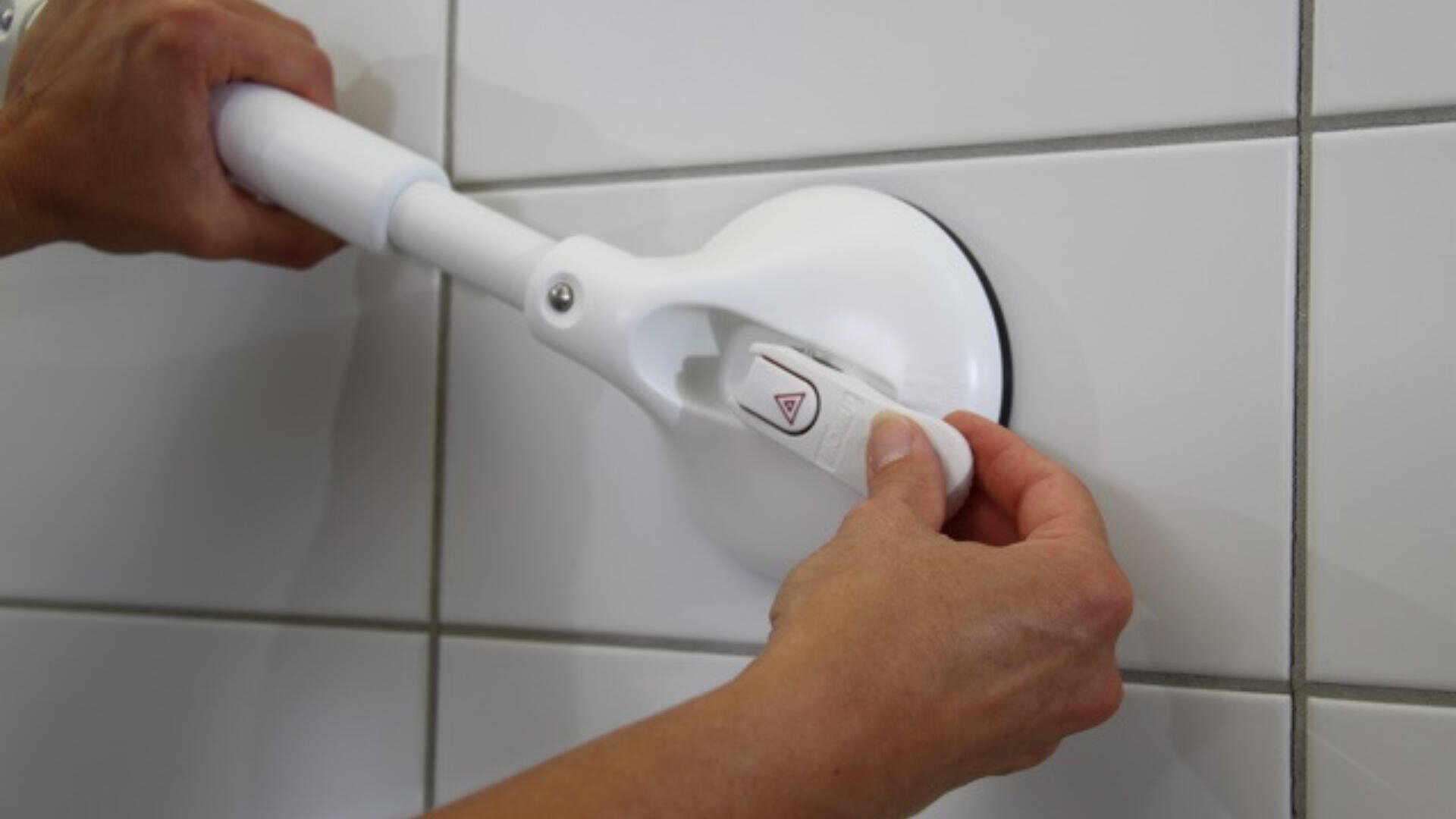 Eine Person befestigt einen weißen Sauggriff an einer gefliesten Wand, indem sie einen Verriegelungshebel nach unten drückt, um Stabilität und Sicherheit zu gewährleisten.