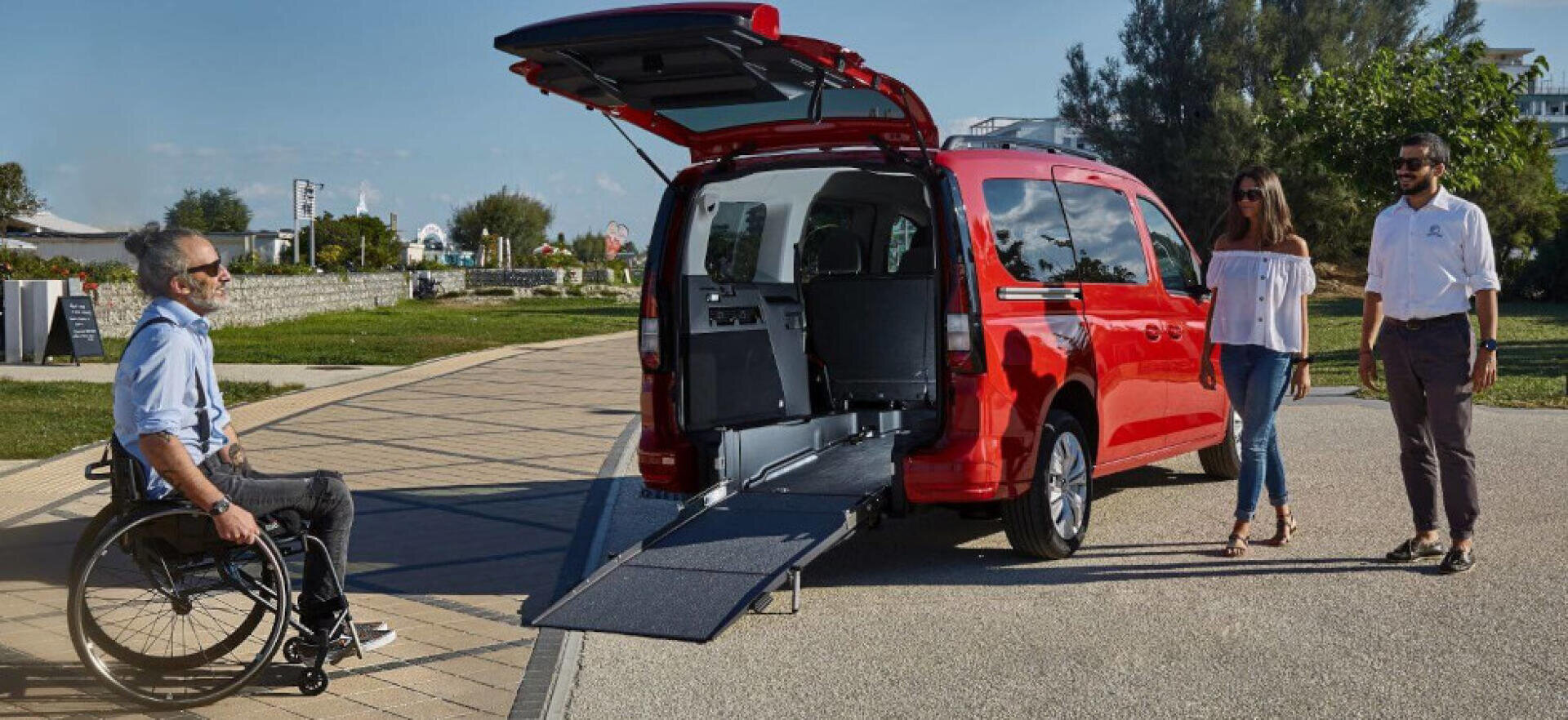 Ein Mann im Rollstuhl nähert sich einem roten Lieferwagen mit offener Heckklappe und ausgefahrener Rampe, während eine Frau und ein Mann an einem sonnigen Tag in einem Park in der Nähe stehen.