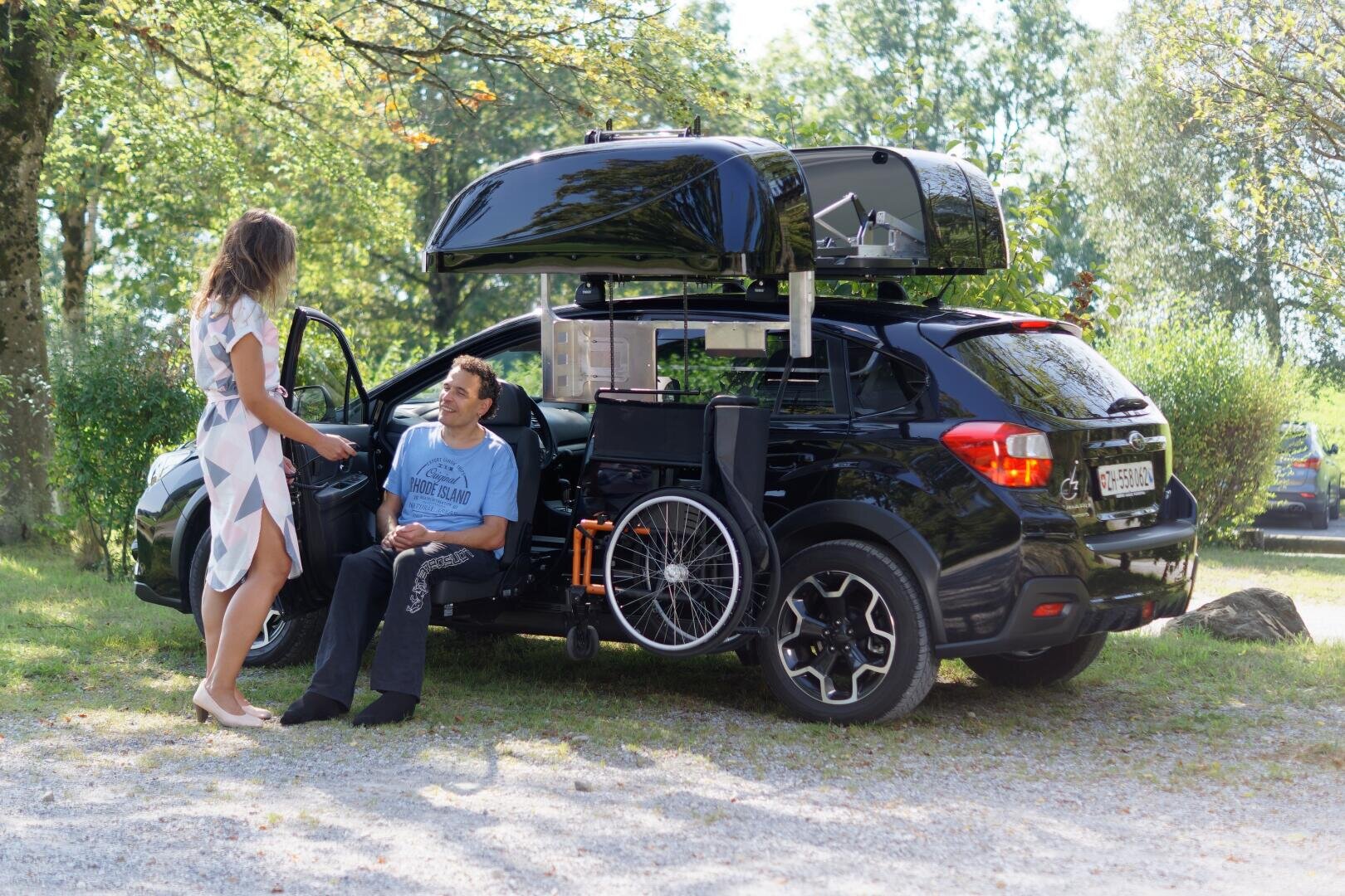 Ein Mann im Rollstuhl sitzt neben einem schwarzen Auto mit offener Dachbox und Seitentür. Eine Frau steht in der Nähe, lächelt und spricht mit ihm. Das Auto ist auf einer Wiese geparkt, mit Bäumen im Hintergrund.