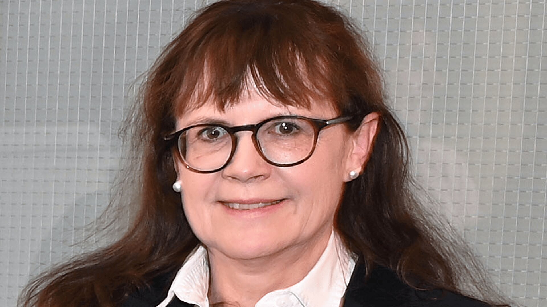 Eine Frau mit langen braunen Haaren und Brille, die ein weißes Hemd mit Kragen, einen dunklen Blazer und Perlenohrringe trägt, lächelt vor einem Hintergrund aus strukturiertem Glas.