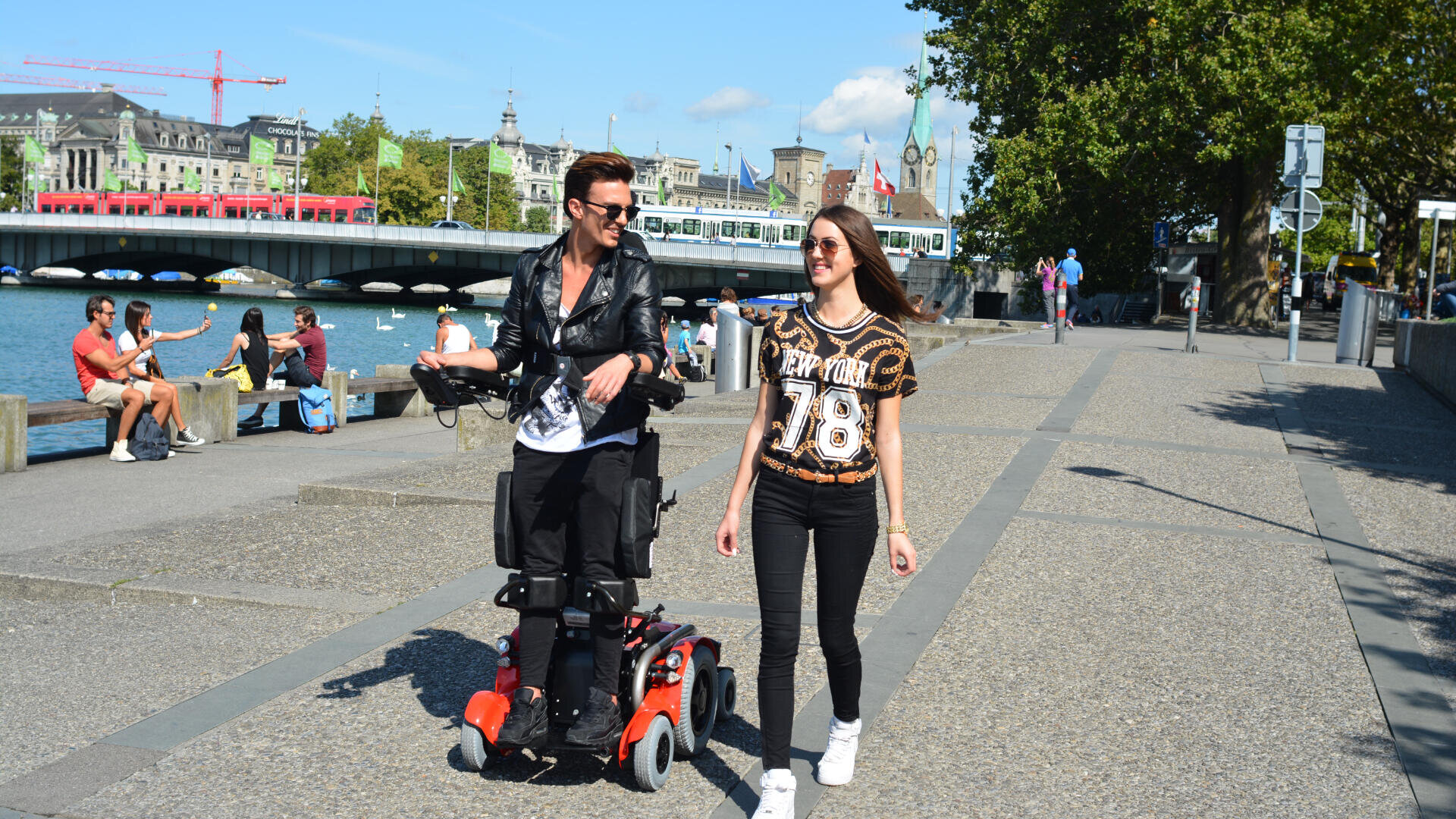 Ein Mann, der einen motorisierten Rollstuhl benutzt, und eine Frau gehen an einem sonnigen Tag gemeinsam lächelnd eine Uferpromenade entlang, mit Menschen, die in der Nähe sitzen, und städtischen Gebäuden im Hintergrund.
