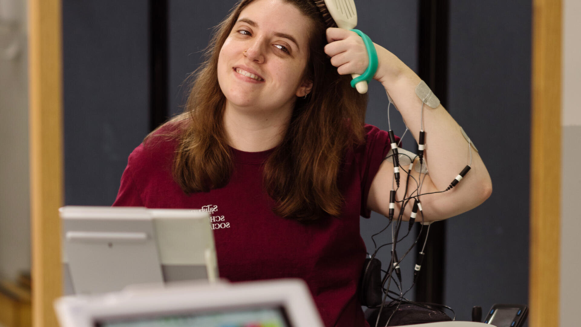 Eine Frau mit langen braunen Haaren benutzt eine weiße adaptive Haarbürste, die an ihrer Hand befestigt ist, mit Elektroden und Drähten an ihrem Arm. Sie lächelt und sitzt vor einem Computerbildschirm in einer Laborumgebung.