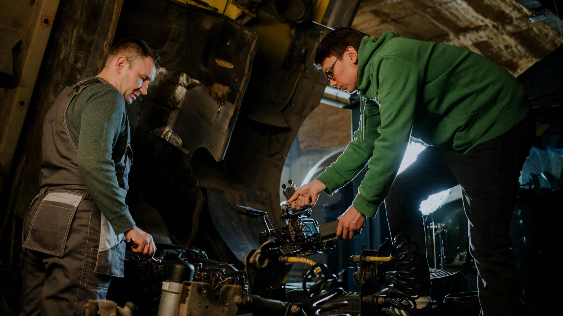 Zwei Männer arbeiten in einer schwach beleuchteten Werkstatt gemeinsam an Maschinen. Einer trägt einen grünen Kapuzenpulli und eine Brille, der andere einen Arbeitsoverall. Sie scheinen sich auf die Reparatur oder Prüfung von Motorenteilen zu konzentrieren.
