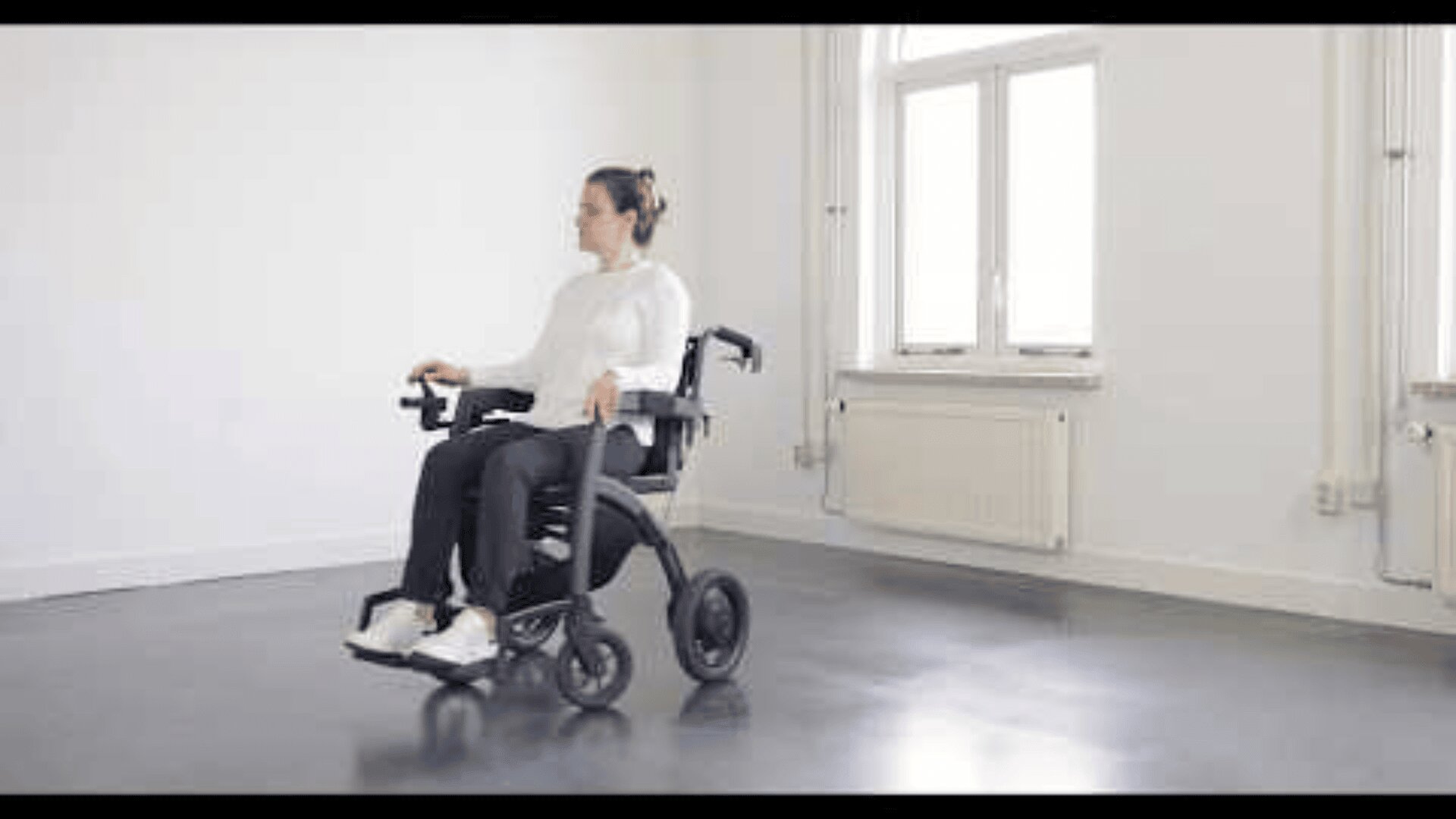 Eine Person in weißem Pullover und schwarzer Hose sitzt in einem modernen elektrischen Rollstuhl in einem hellen, leeren Raum mit großen Fenstern und grauem Fußboden.