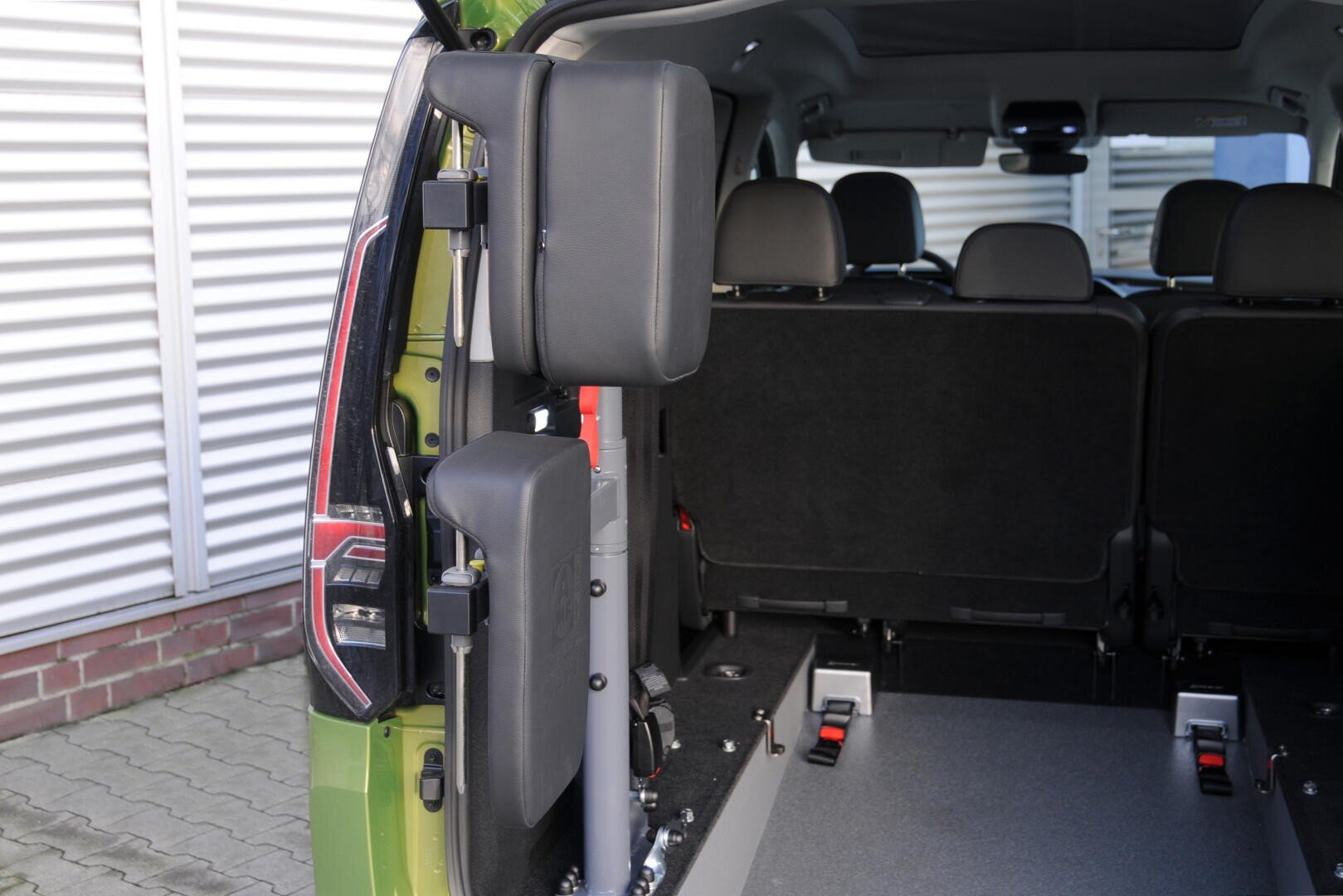 Das Bild zeigt den hinteren Innenraum eines Lieferwagens, der mit einem zusammengeklappten Rollstuhllift in der Nähe der offenen Hintertür ausgestattet ist. Im Inneren des Fahrzeugs sind Sitze und Befestigungsschienen zu sehen.