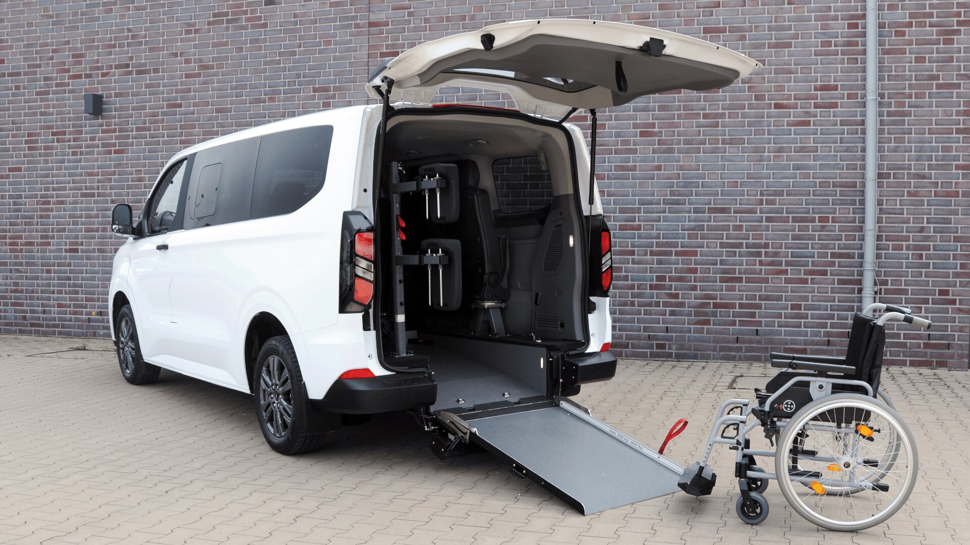 Ein weißer Lieferwagen mit geöffneter Hecktür und einer bis zum Boden ausgefahrenen Rampe, der neben einem leeren Rollstuhl auf einer gepflasterten Fläche steht, zeigt die Zugänglichkeitsmerkmale.