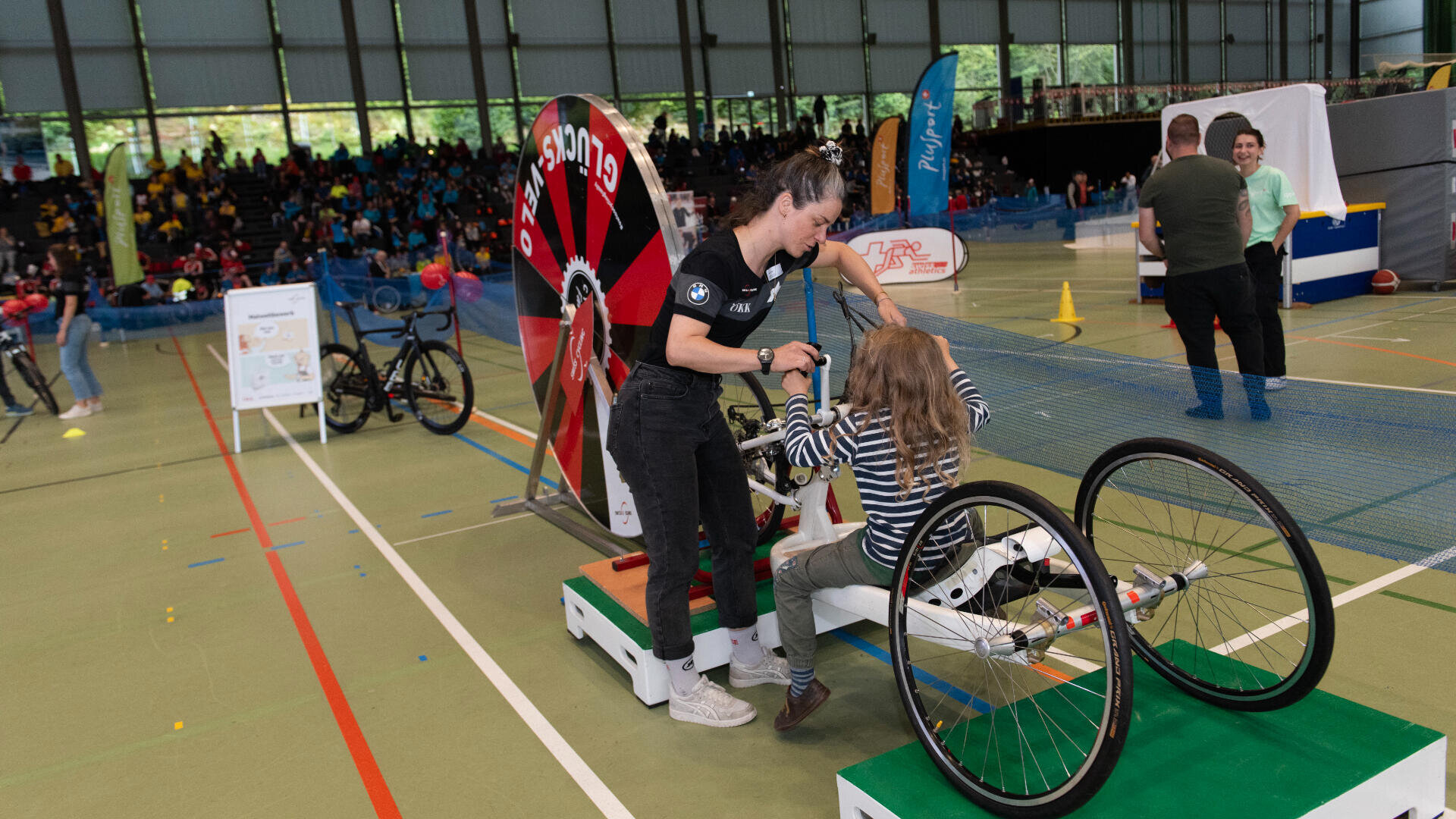 Eine Frau hilft einem Mädchen, sich während einer Sportveranstaltung in einer großen Sporthalle auf einen Handbike-Simulator zu setzen, wobei im Hintergrund Menschen, Fahrräder und bunte Banner zu sehen sind.