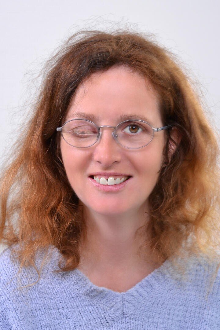 Eine Frau mit gewelltem, hellbraunem Haar, die eine Brille und einen hellblauen Pullover trägt, lächelt mit einem geschlossenen und einem offenen Auge in die Kamera. Der Hintergrund ist einfarbig und hell.