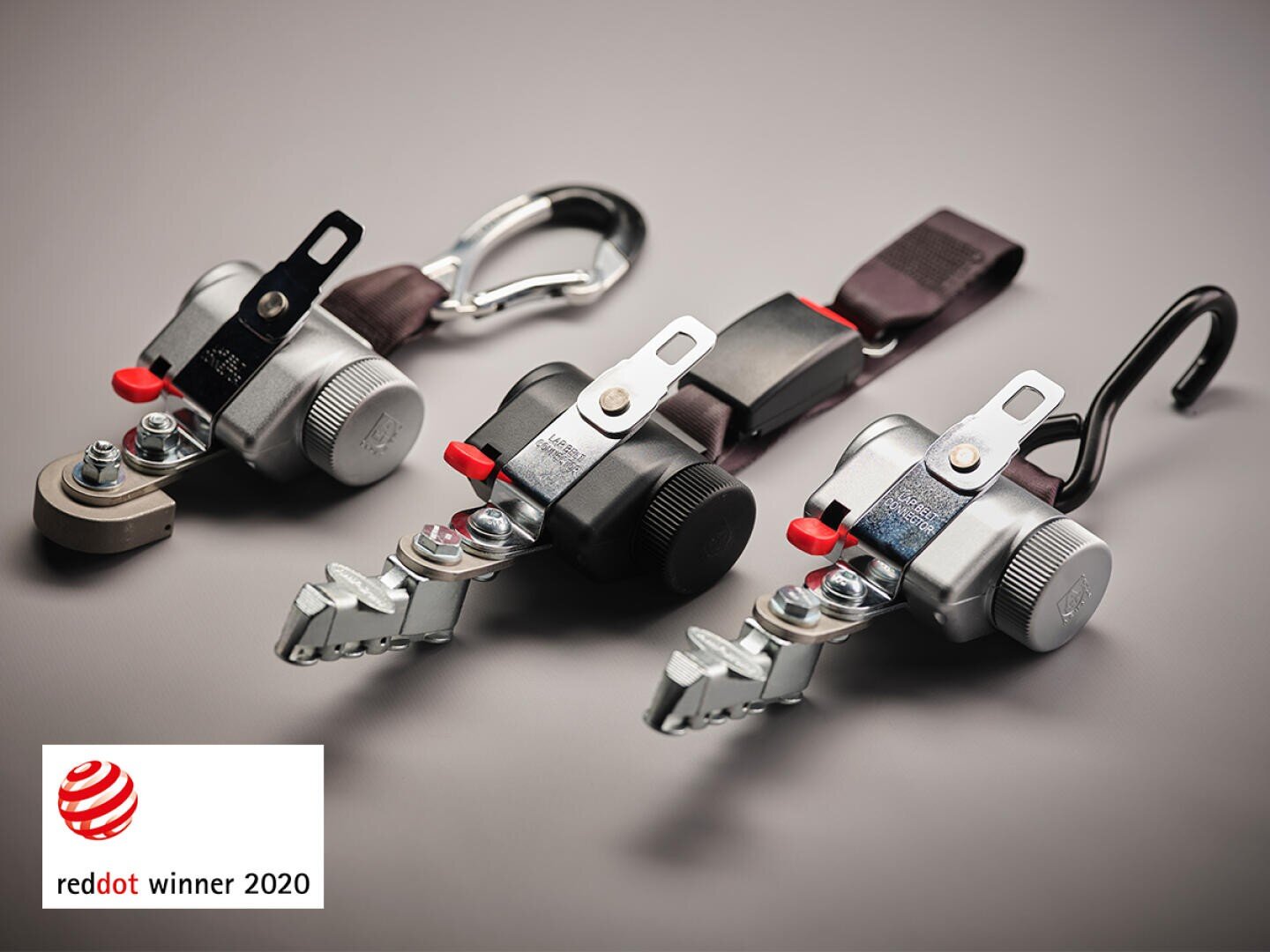 Drei schwarze und silberne mechanische Vorrichtungen mit Haken und Verschlüssen sind auf einer grauen Fläche abgebildet. In der unteren linken Ecke erscheint das Logo des Red Dot Winner 2020.