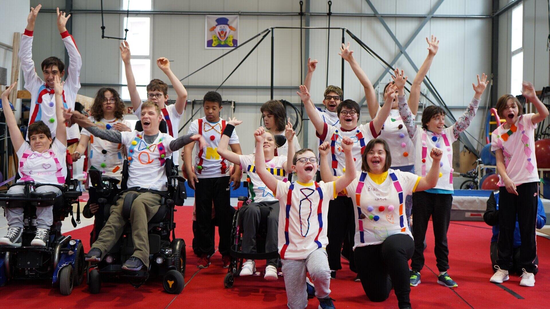Eine bunt gemischte Gruppe von Kindern, von denen einige im Rollstuhl sitzen, feiert gemeinsam auf einem roten Teppich in einer Halle. Sie tragen farbenfrohe, gemusterte Hemden und haben die Arme hochgehoben, lächeln und jubeln enthusiastisch.