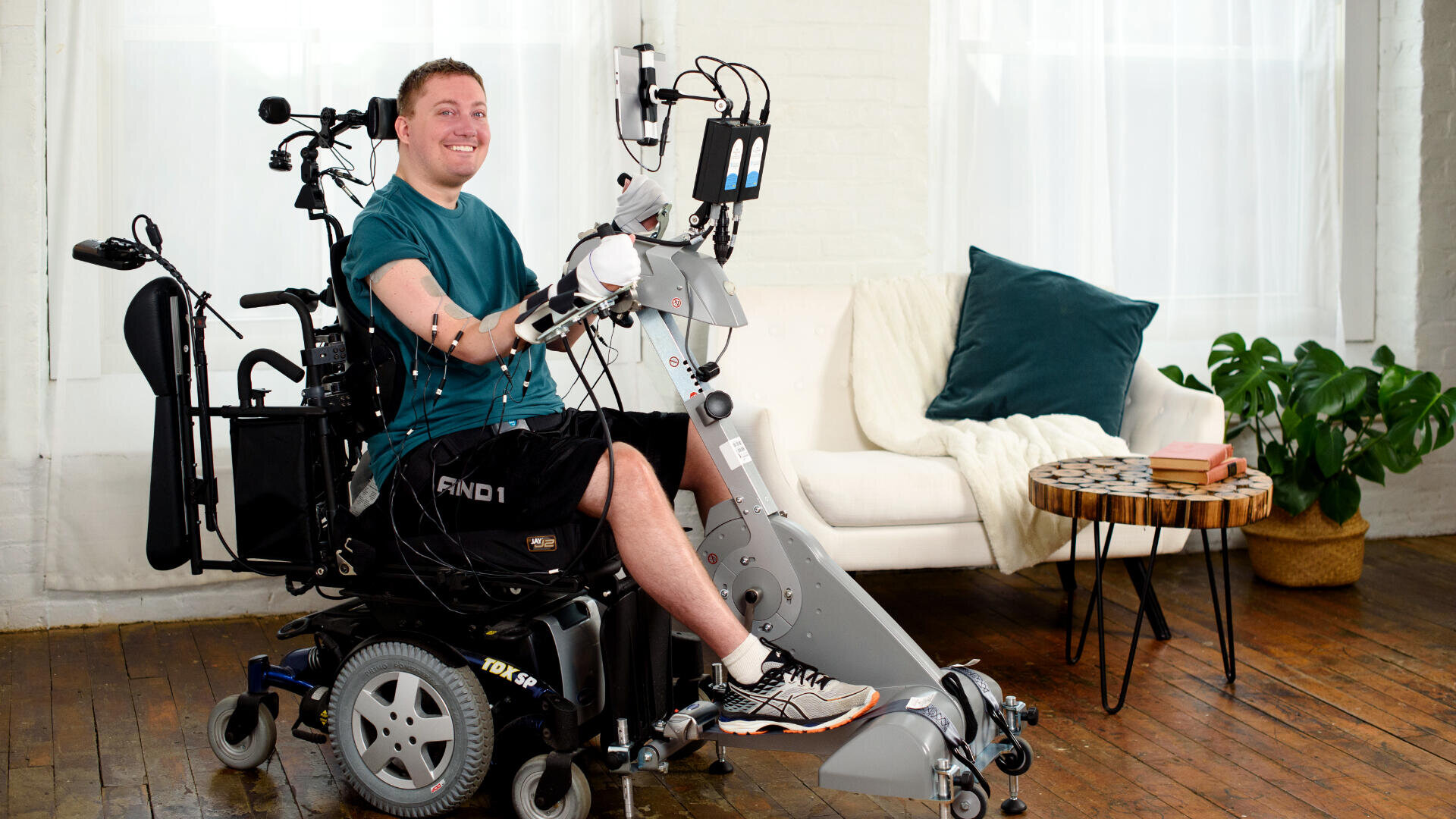 Ein lächelnder Mann im Rollstuhl benutzt einen Roboterarm, der mit Drähten an seinen Armen befestigt ist. Er sitzt in einem Innenraum auf einem Holzboden, neben einem weißen Sofa mit blaugrünen Kissen, einem runden Tisch und einer grünen Pflanze.