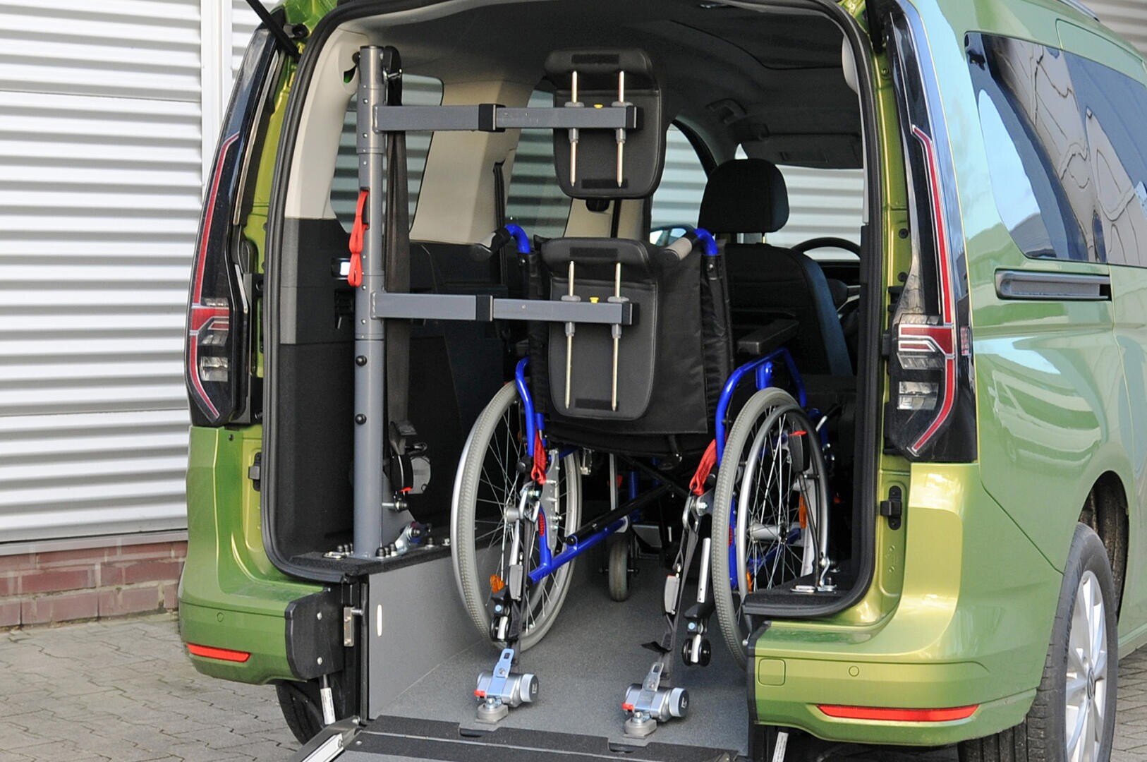 Ein grüner Lieferwagen mit geöffneter Hecktür, in dem ein Rollstuhl sicher auf einer Rampe befestigt ist, was darauf hinweist, dass das Fahrzeug für den Zugang von Rollstuhlfahrern ausgerüstet ist.