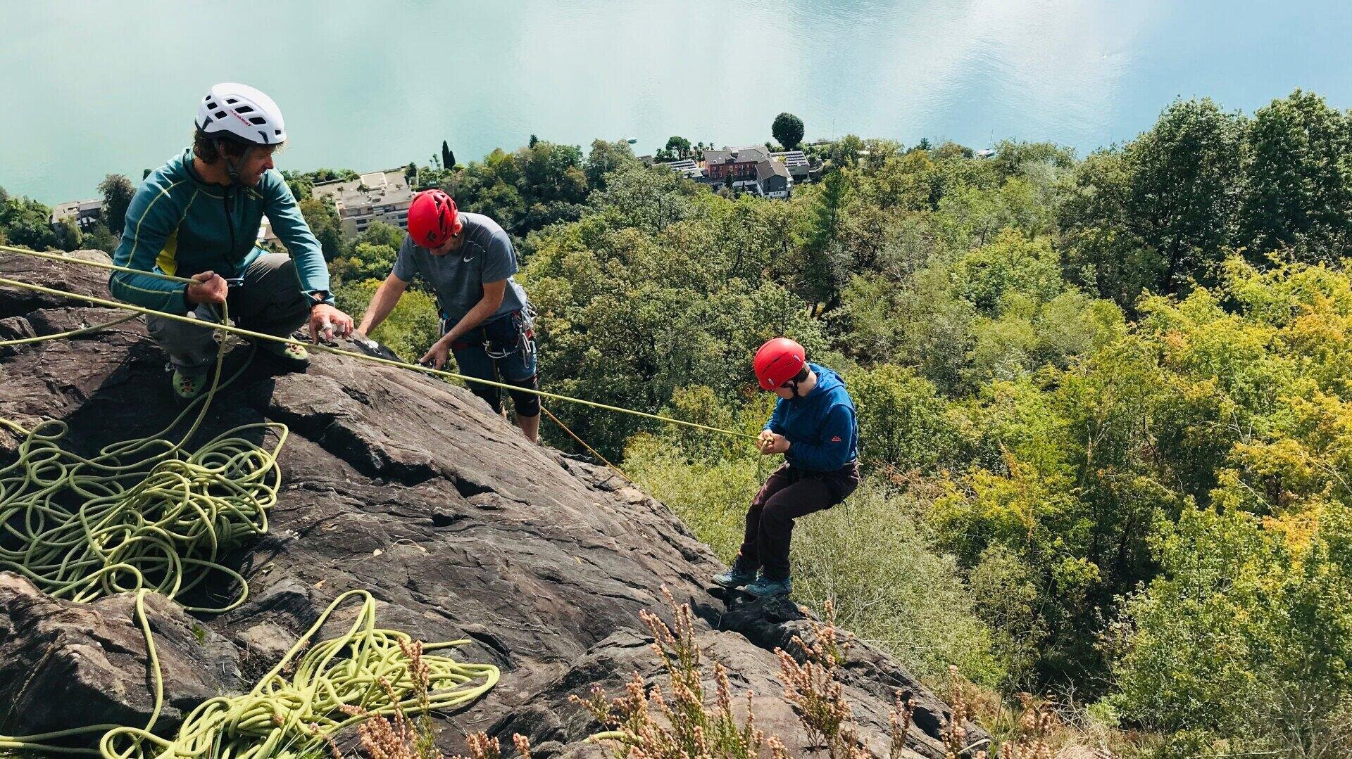 Drei Personen mit Helmen und Kletterausrüstung klettern an einem steilen Felsen, wobei sie Seile zur Sicherung benutzen. Im Hintergrund sind Bäume und ein Gewässer zu sehen.