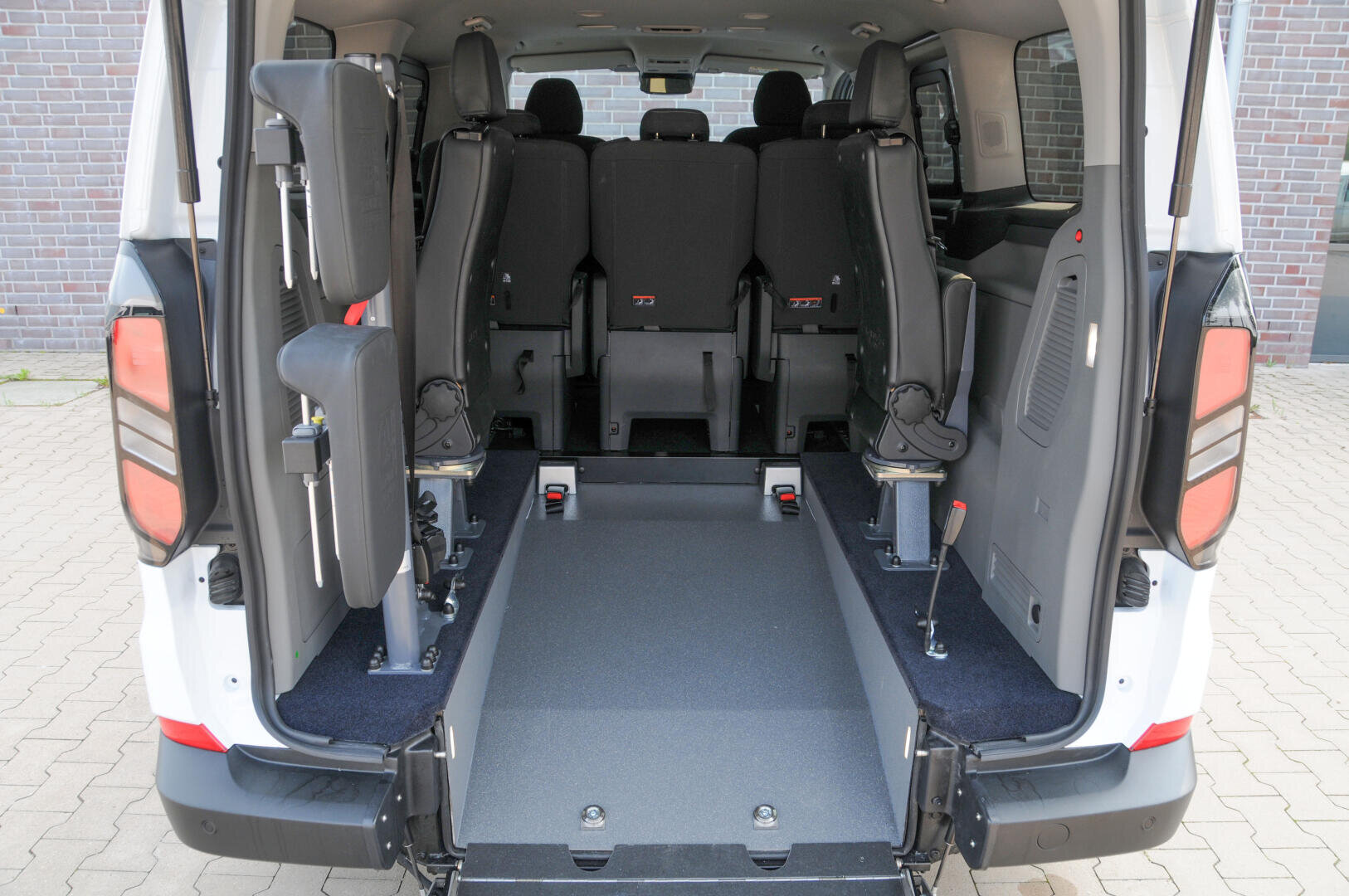 Das Bild zeigt die Rückansicht eines rollstuhlgerechten Lieferwagens mit geöffneten Hintertüren, einem abgesenkten Boden und umgeklappten Sitzen an den Seiten, damit ein Rollstuhl im Mittelgang Platz findet.