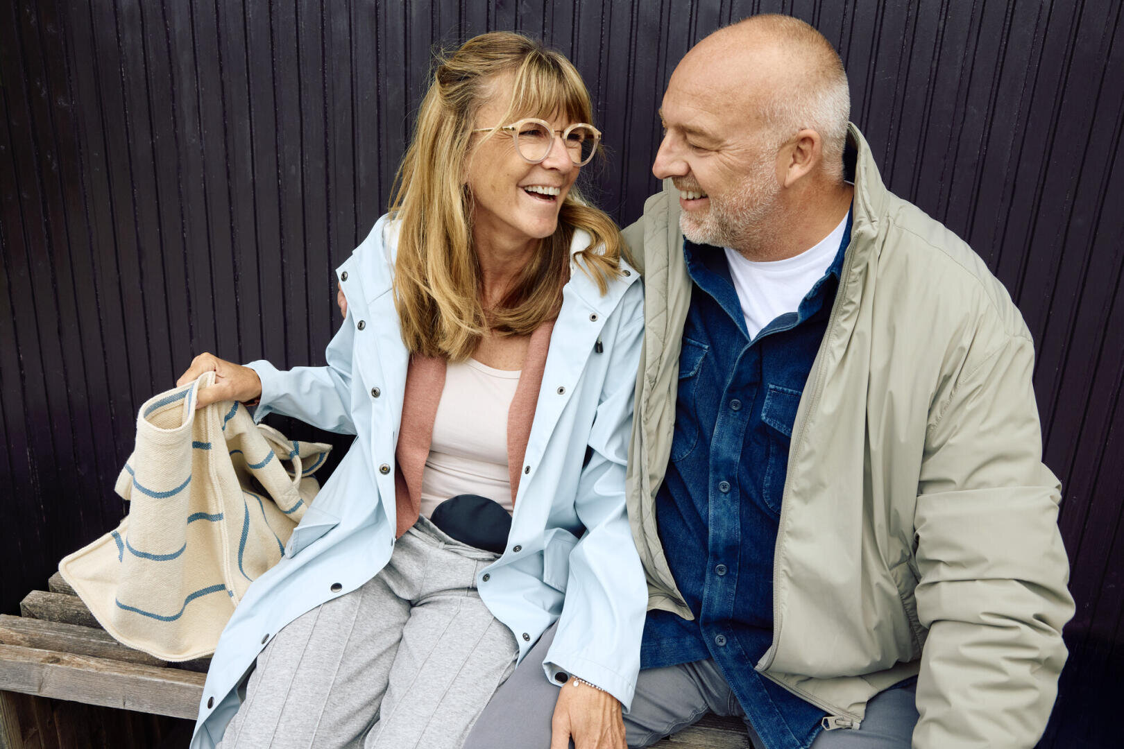 Ein lächelndes älteres Paar sitzt eng beieinander auf einer Bank und trägt legere Jacken. Die Frau hält eine gestreifte Tasche in der Hand, und die beiden schauen sich glücklich an. Sie teilen einen freudigen Moment vor einem dunklen Holzhintergrund.