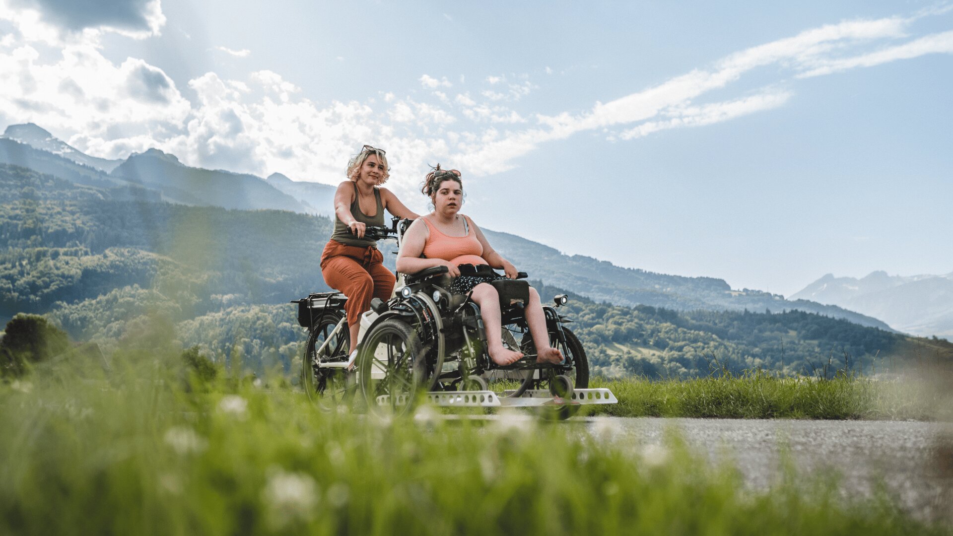 Zwei Frauen fahren mit einem an den Rollstuhl angepassten Fahrrad auf einem Weg durch grüne Felder, mit Bergen und einem bewölkten Himmel im Hintergrund. Eine Frau tritt in die Pedale, während die andere vorne sitzt.