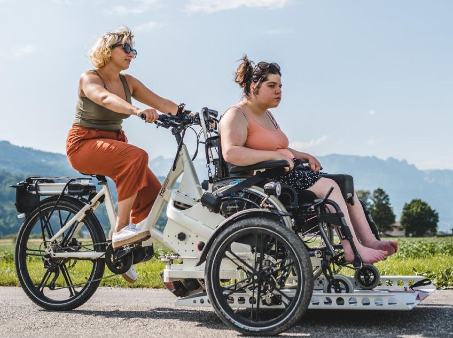 Zwei Frauen im Freien: Die eine fährt auf einem umgebauten Fahrrad, das die andere, die in einem Rollstuhl sitzt, sicher festhält. Sie befinden sich auf einem gepflasterten Weg, mit Bergen und Bäumen im Hintergrund an einem sonnigen Tag.