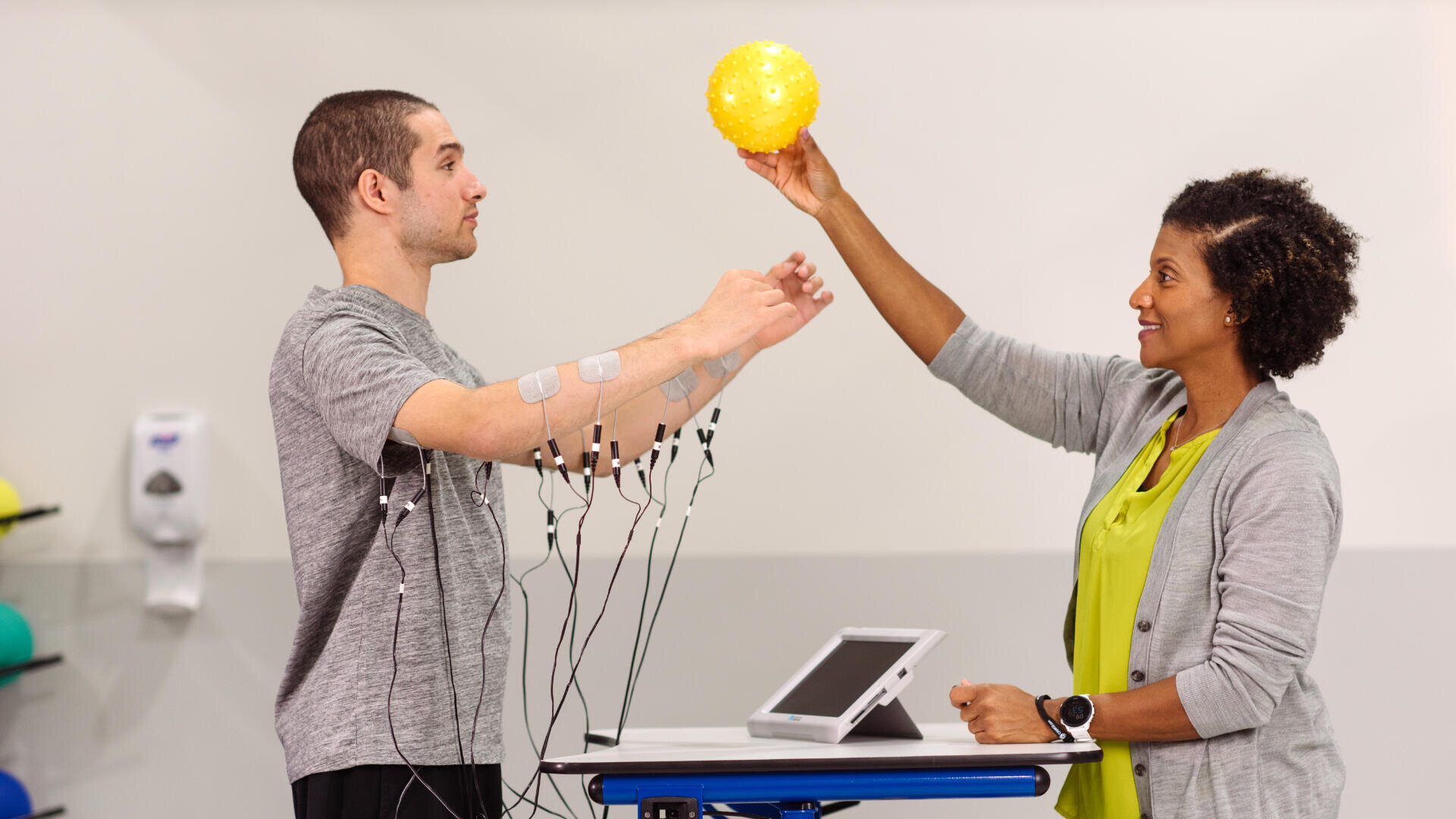 Eine Frau hält einen gelben Ball über die ausgestreckten Arme eines Mannes. Der Mann hat Elektroden an seinen Armen, die mit einem Gerät auf einem Tisch verbunden sind. Sie scheinen sich in einer Physiotherapie- oder Rehabilitationseinrichtung zu befinden.