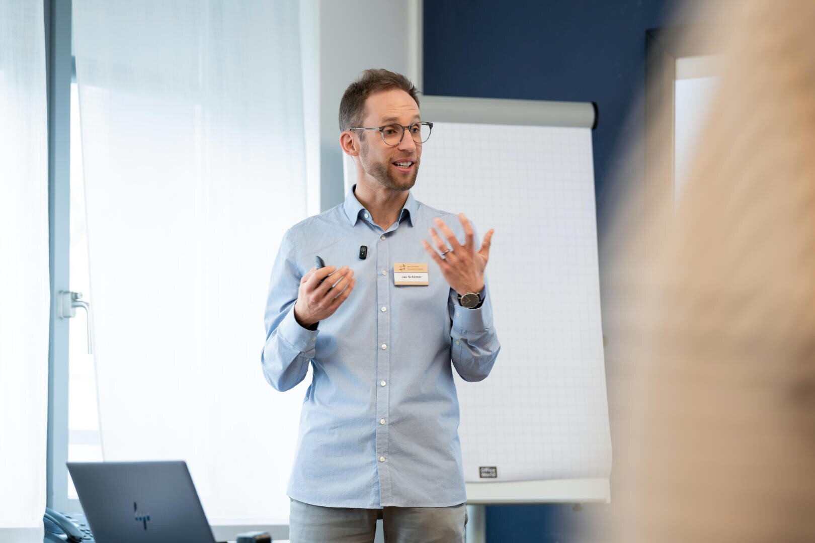 Ein Mann mit Brille und in einem hellblauen Hemd steht vor einem Flipchart, spricht und gestikuliert dabei mit einer Hand. Auf dem Tisch daneben steht ein Laptop, und durch ein Fenster fällt Tageslicht herein.