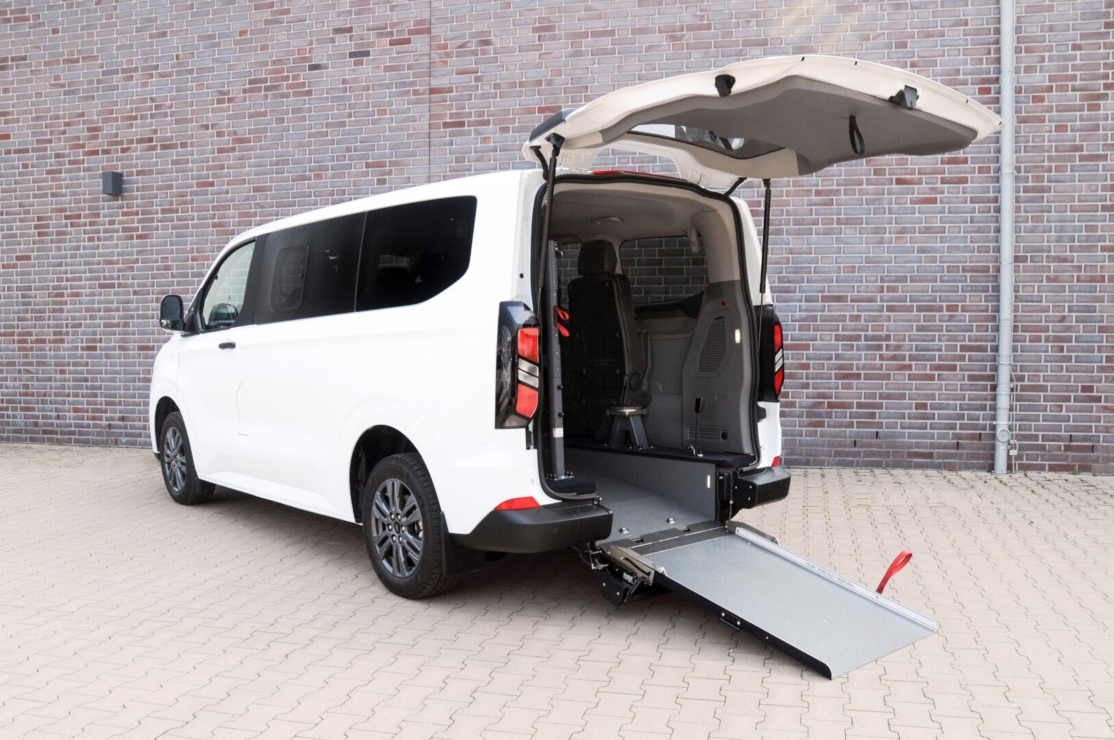 Ein weißer Lieferwagen mit geöffneter Hecktür, die eine Rampe zeigt, die für den Zugang mit dem Rollstuhl auf den Boden ausgefahren ist. Der Lieferwagen ist auf einer gepflasterten Fläche vor einer Backsteinmauer geparkt.