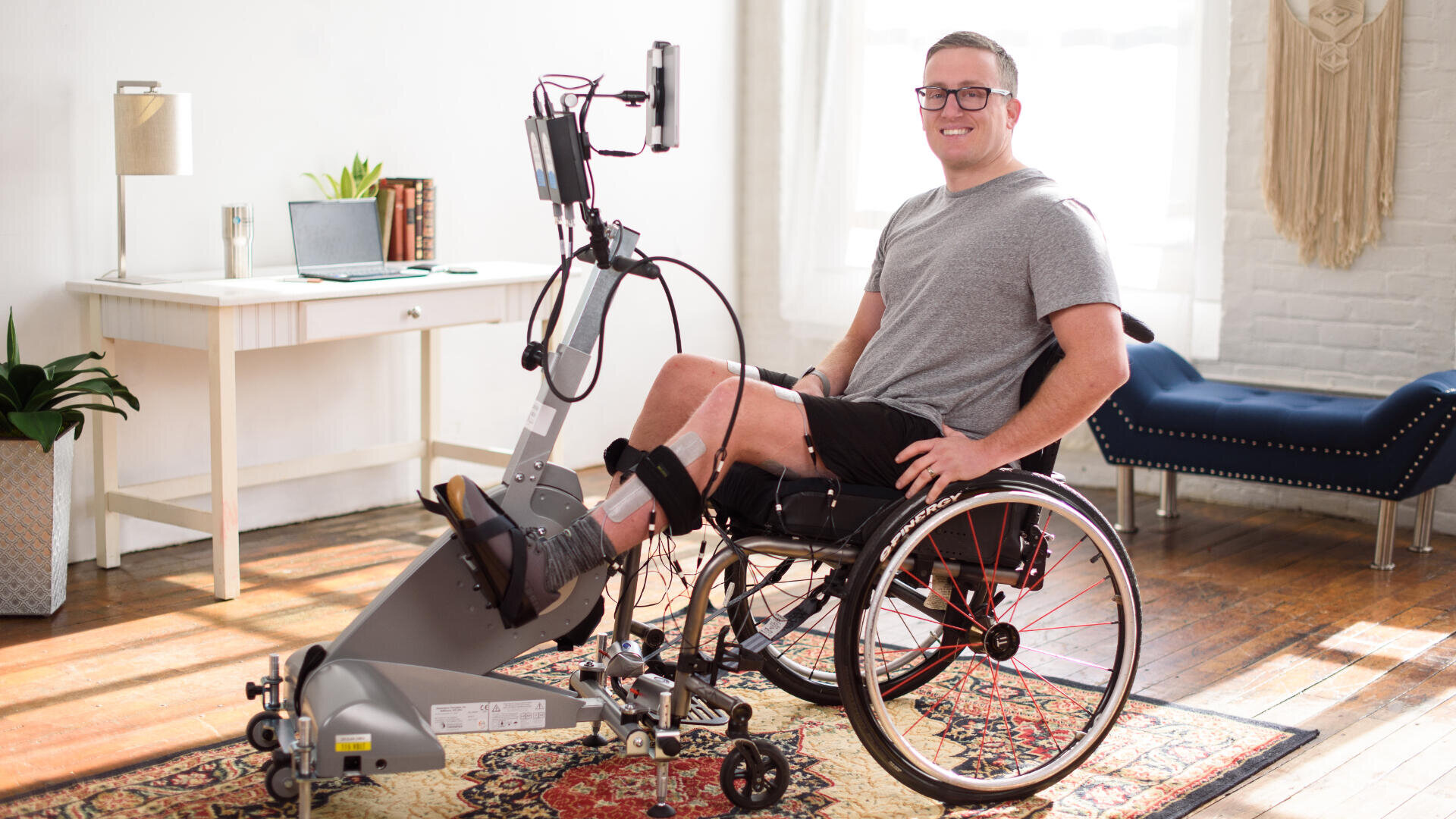 Ein Mann im Rollstuhl benutzt ein Beintrainingsgerät in einem hellen, wohnlich eingerichteten Raum. Er lächelt, trägt eine Brille und ein graues Hemd. Im Hintergrund sind ein Schreibtisch, Pflanzen und ein Fenster mit natürlichem Licht zu sehen.