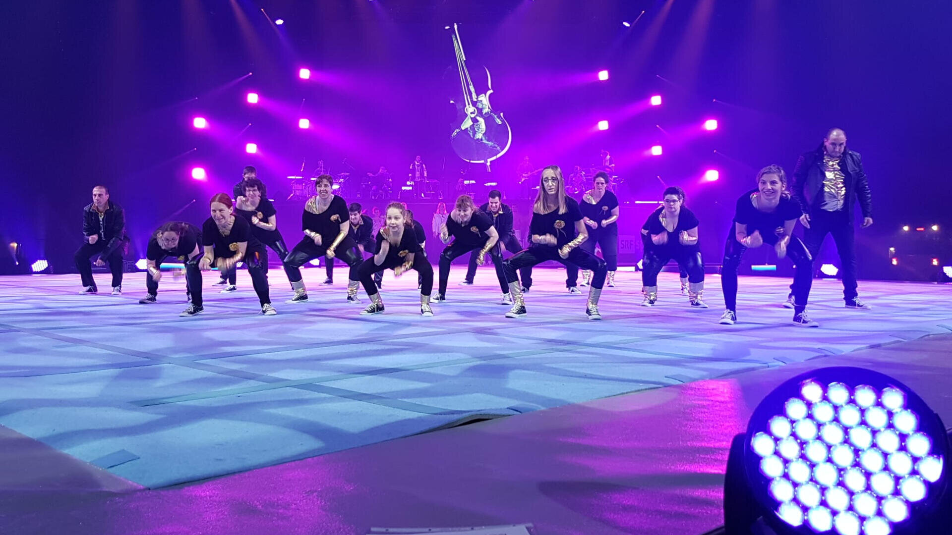Eine schwarz gekleidete Tanzgruppe tritt energisch auf einer mit violetten Lichtern beleuchteten Bühne auf, während im Hintergrund Musiker und eine große Gitarrendekoration zu sehen sind.