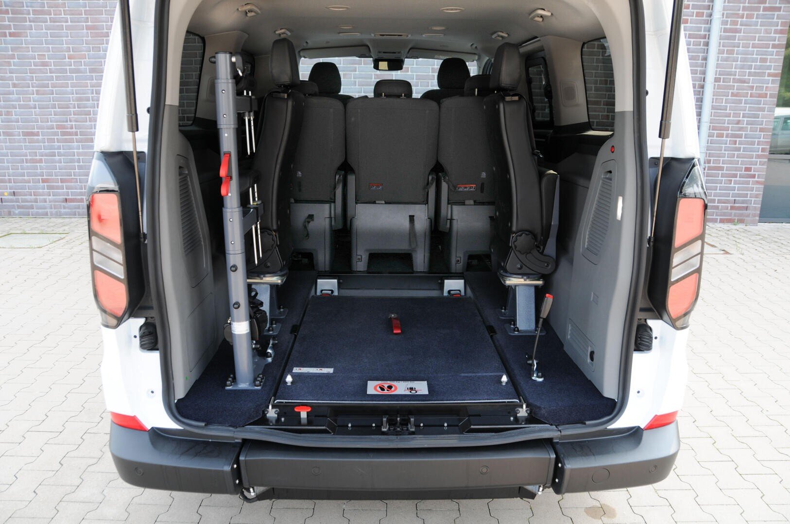 Das Bild zeigt die Rückansicht eines Lieferwagens mit geöffneten Hintertüren, die den Blick auf einen geräumigen Innenraum mit umgeklappten Sitzen und einer Plattform freigeben, auf der ein Rollstuhl oder eine Mobilitätshilfe untergebracht werden kann.