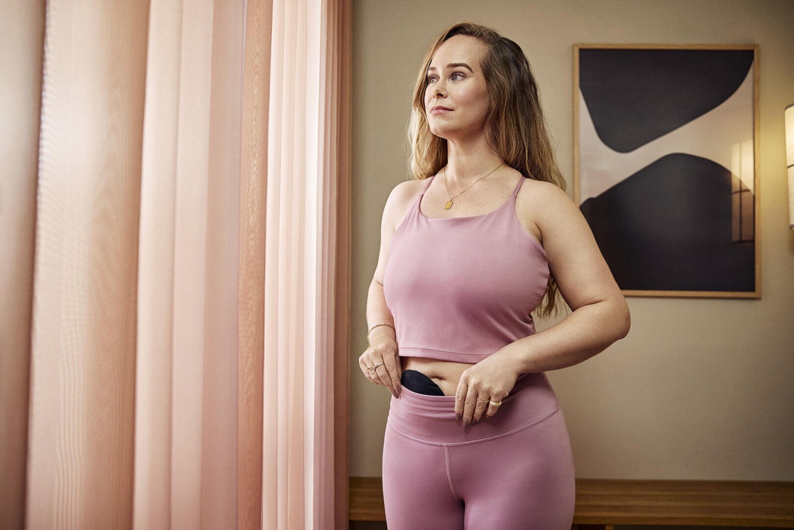 Eine Frau in rosa Sportkleidung steht in einem Raum, hält ein schwarzes Gerät an ihrem Hosenbund fest und blickt nachdenklich aus einem Fenster mit rosa Vorhängen. Im Hintergrund sind moderne Kunstwerke und eine Lampe zu sehen.