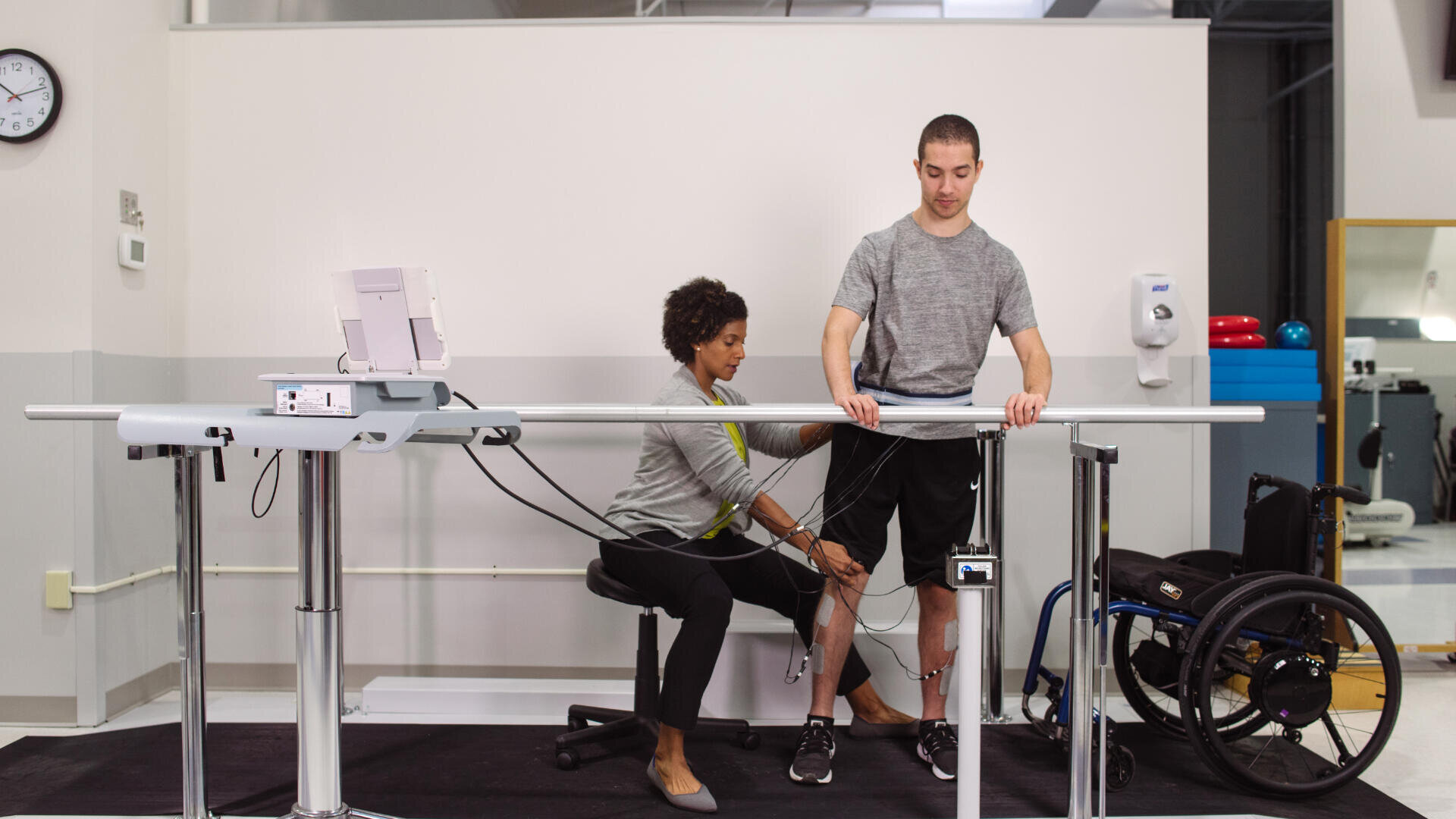 Ein Physiotherapeut bringt Sensoren an den Beinen eines Mannes an, der an einem hohen medizinischen Arbeitsplatz steht; in der Nähe befindet sich ein Rollstuhl, der auf die Rehabilitation in einem klinischen Umfeld hinweist.