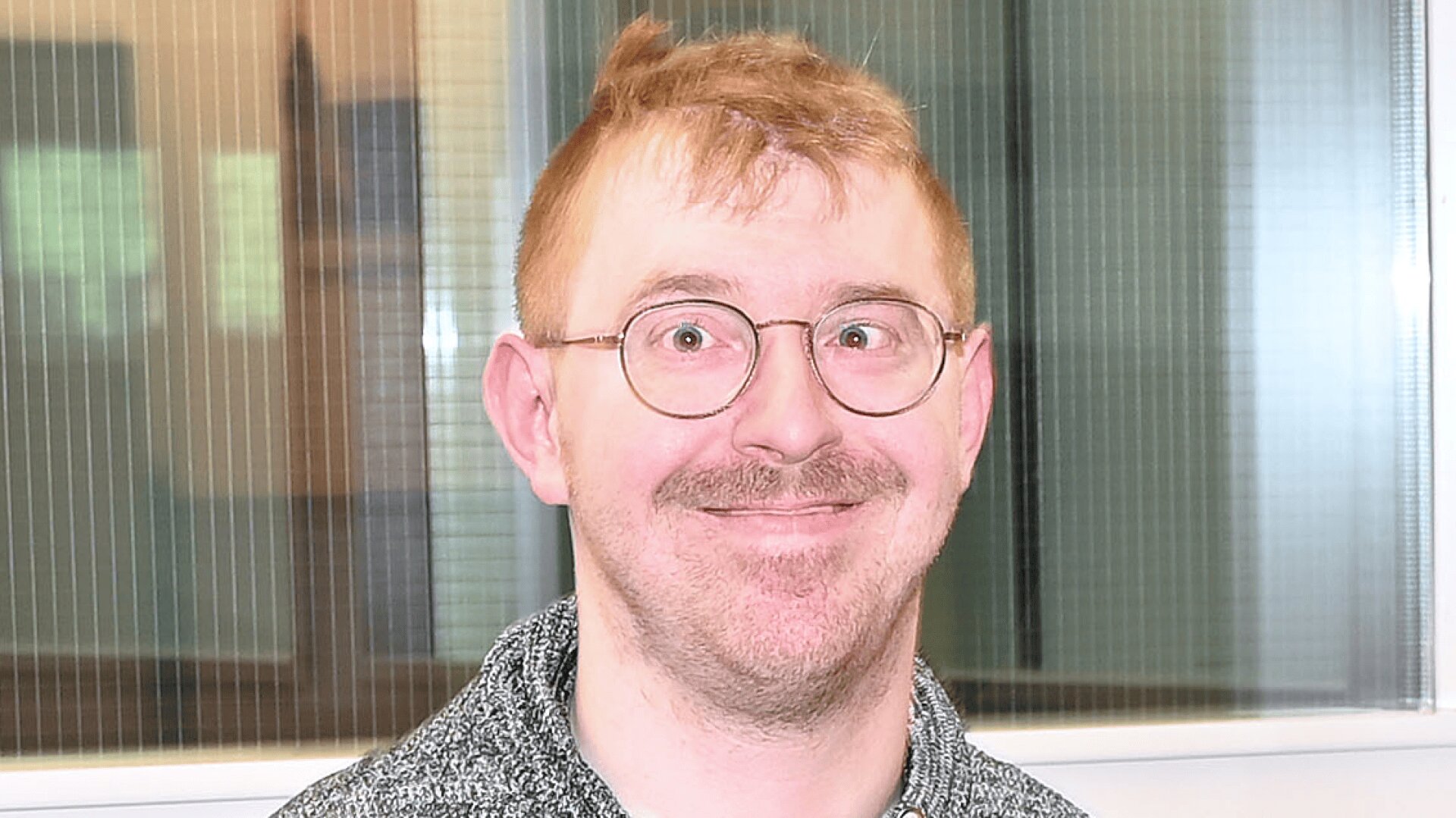 Eine Person mit kurzen, hellen Haaren, einer Brille und einem Schnurrbart steht lächelnd vor einem Fenster mit vertikalen Jalousien. Sie trägt ein graues, gemustertes Oberteil.