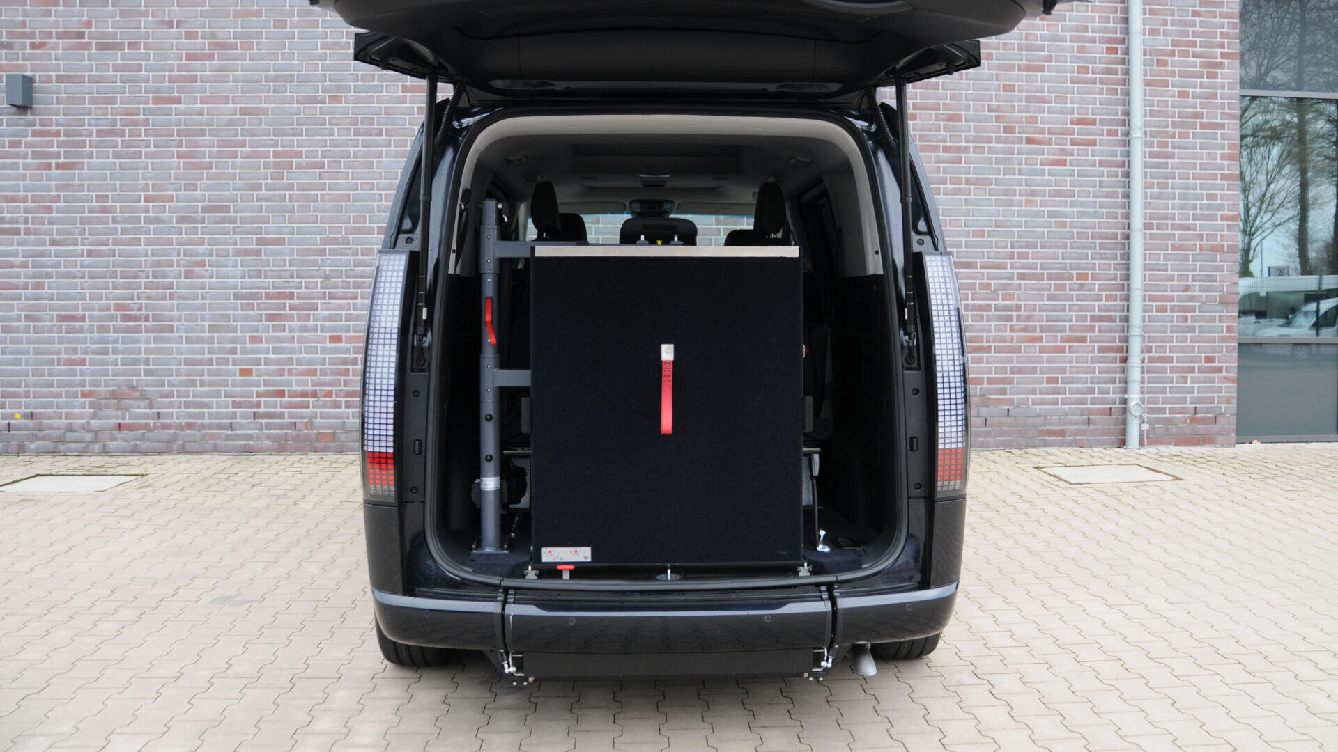 Ein schwarzer Lieferwagen mit geöffneter Heckklappe, in der eine große, aufrecht stehende Plattform für einen Rollstuhllift zu sehen ist. Der Lieferwagen ist auf einer gepflasterten Fläche vor einer Backsteinmauer geparkt.